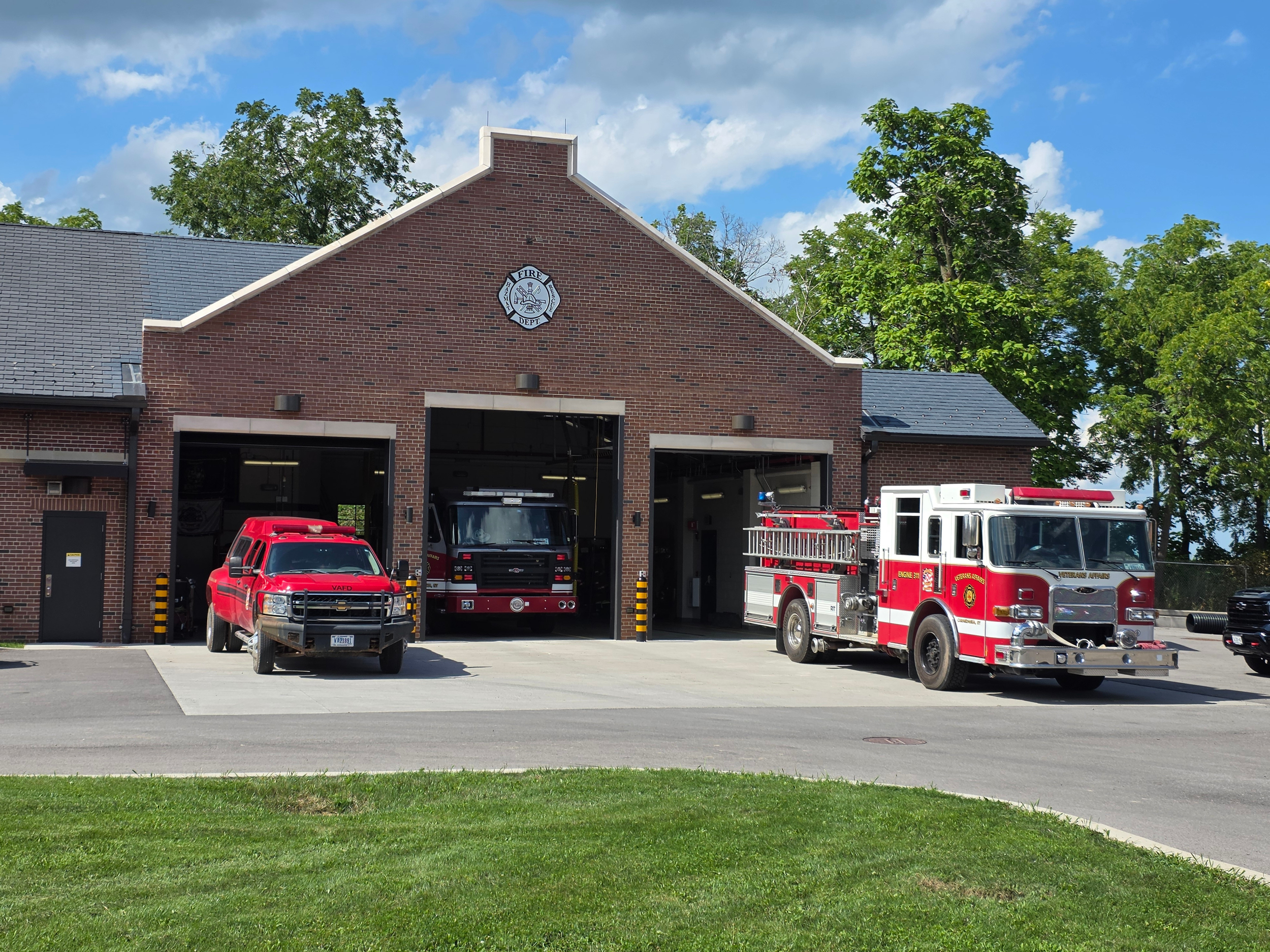 Fire station with three emergency vehicles parked outside under a partly cloudy sky, surrounded by green trees.