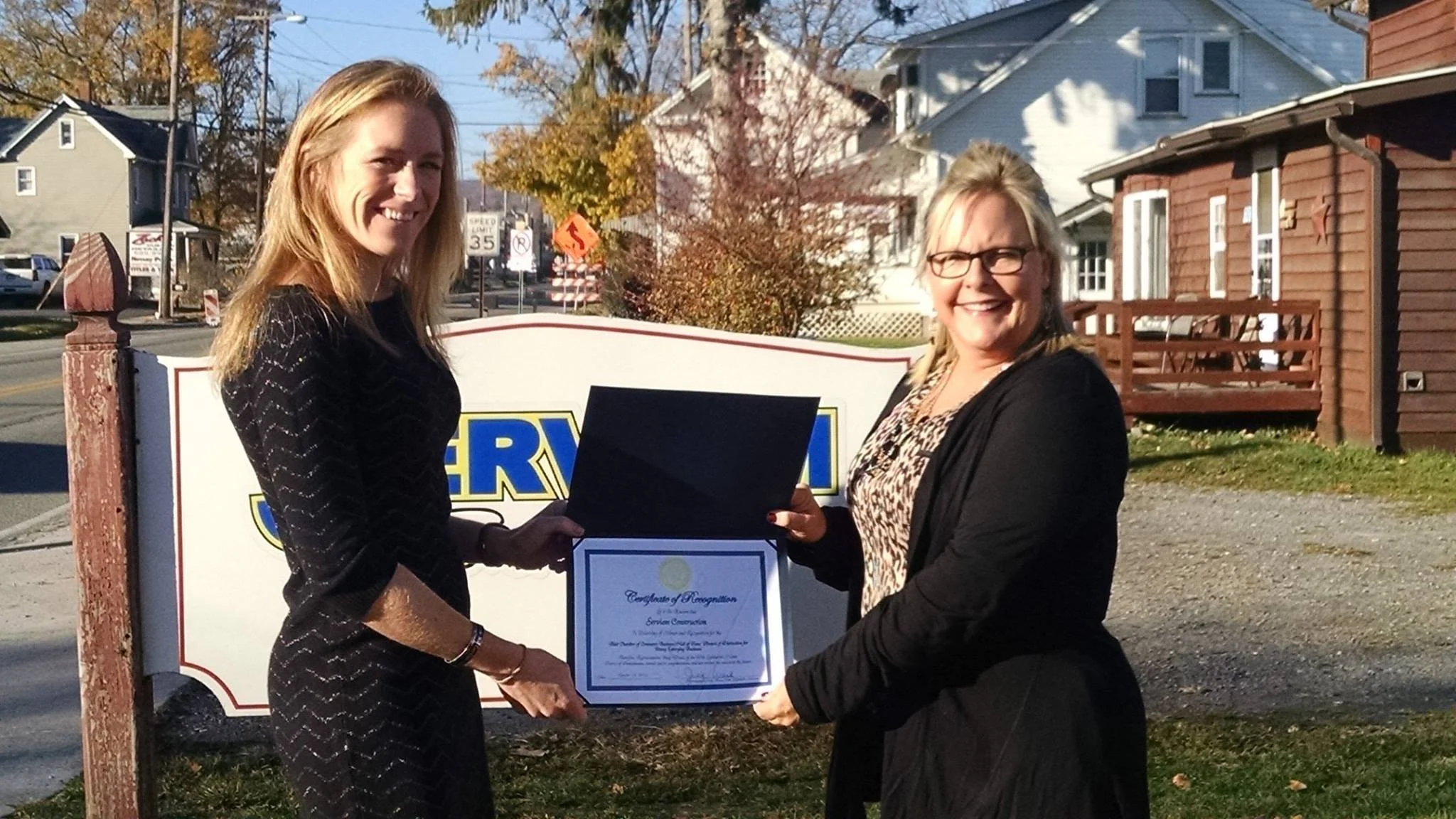 Two women smiling and shaking hands outdoors in front of a sign, one is holding a certificate, during a daytime ceremony or event.