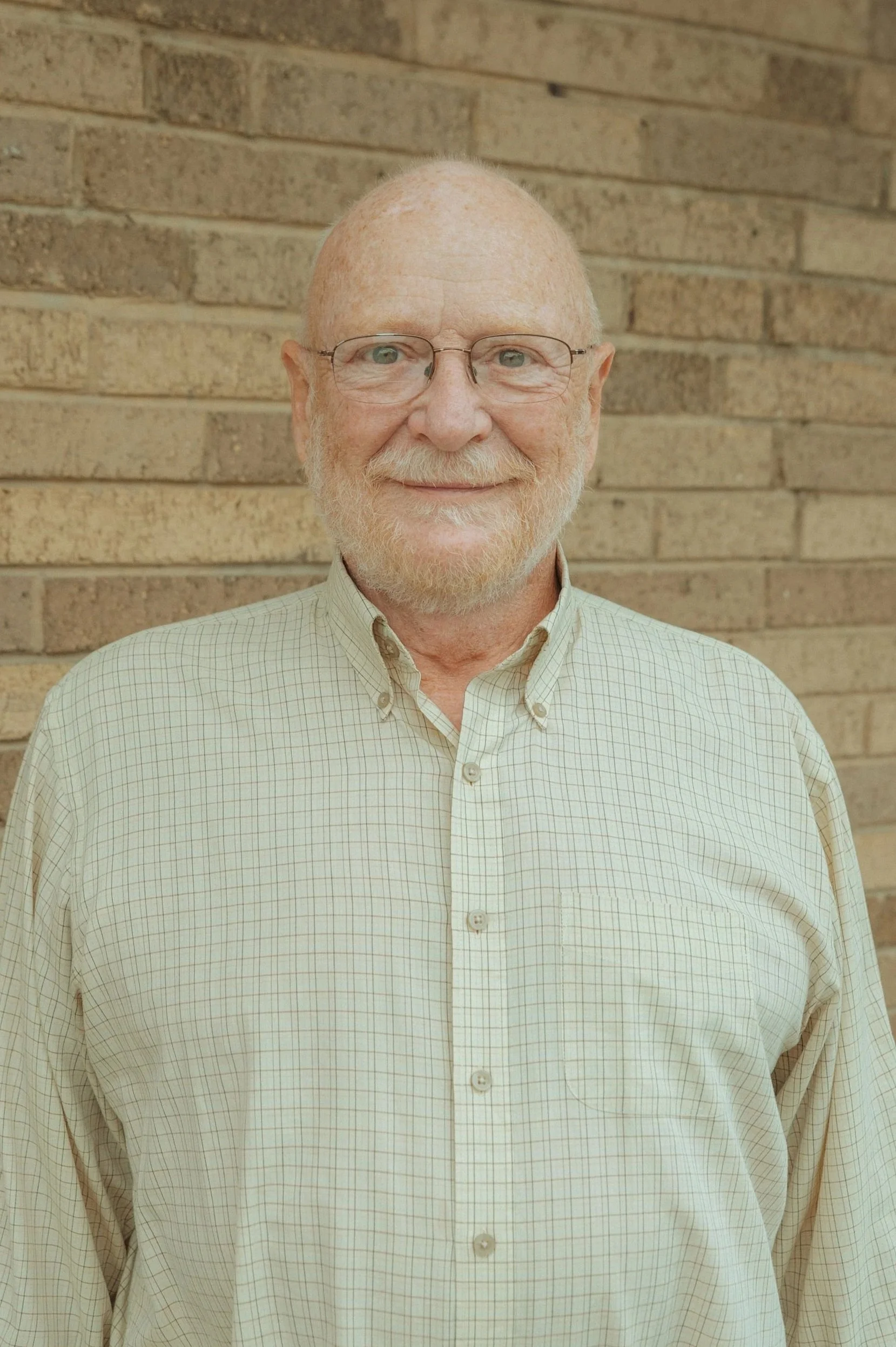 Close-up portrait of an older man with glasses, a beard, and light-colored hair, smiling and wearing a cream-colored checkered button-up shirt, standing against a brick wall.