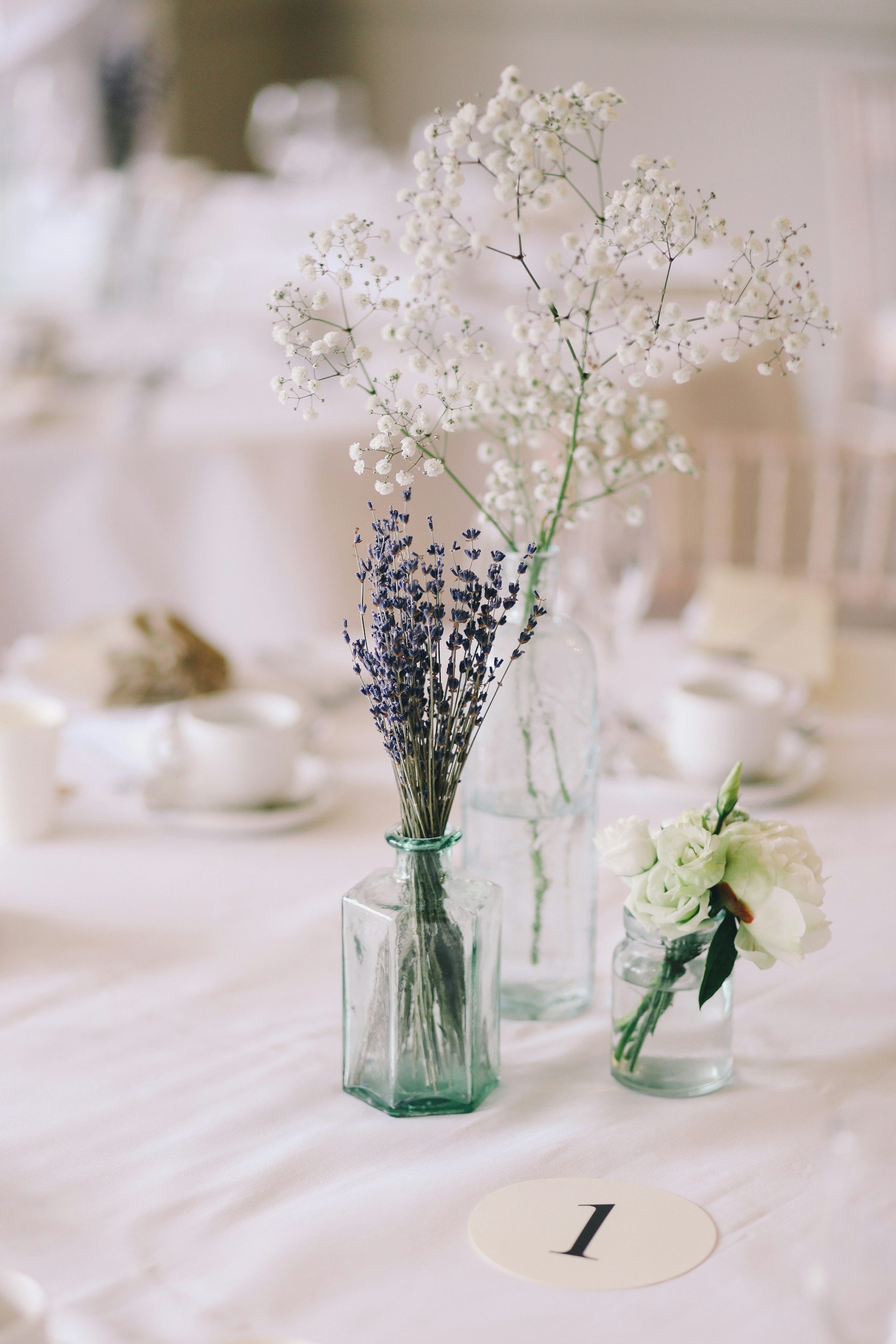A centerpiece with white baby's breath and purple lavender in glass vases on a white tablecloth, with a table number '1' card in front.