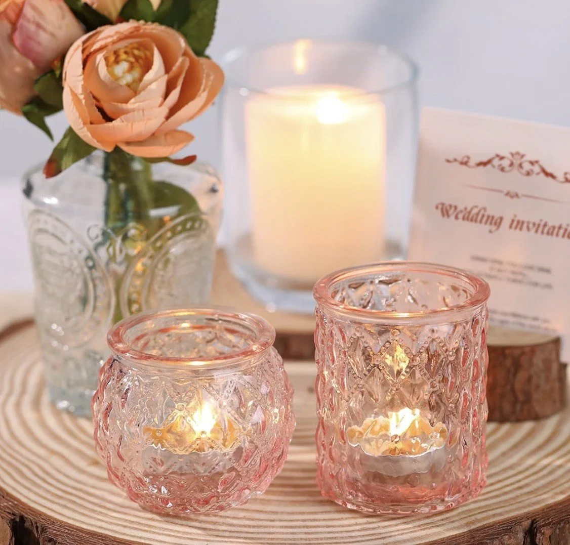 Decorative candles in pink glass holders, a lit white candle in a clear glass container, a bouquet of peach-colored roses in a clear embossed glass vase, and a wedding invitation on a wooden tray, all on a round wooden base.
