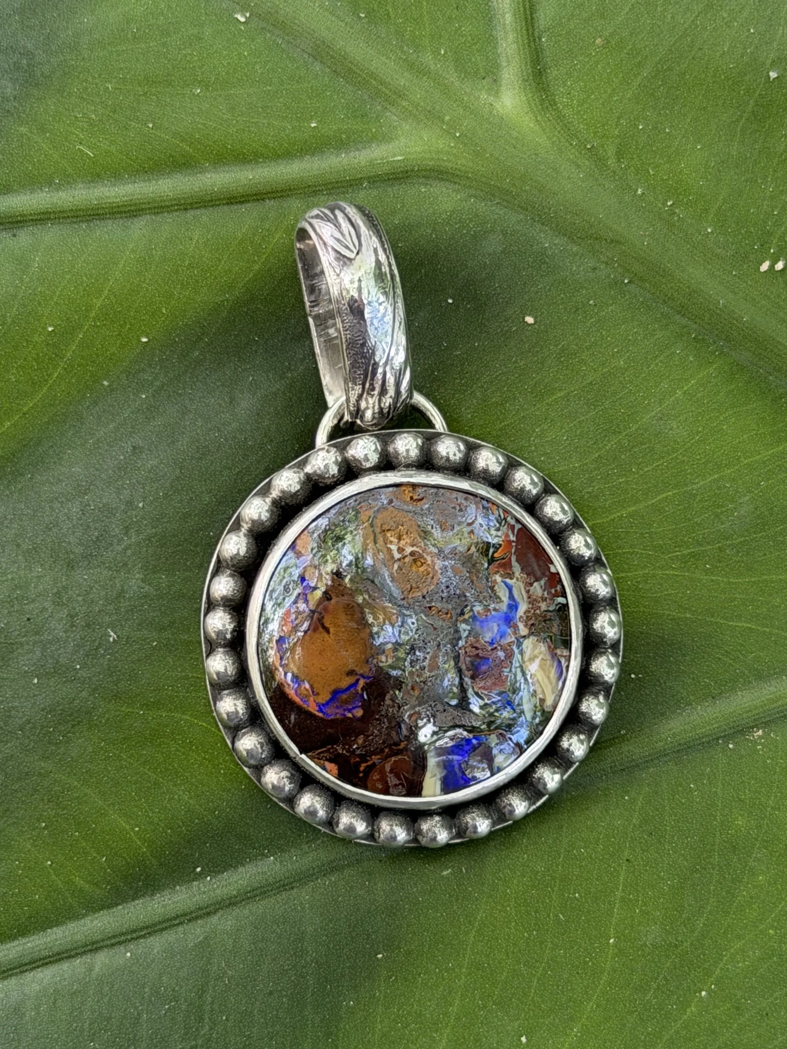 Close-up image of a jewelry pendant with a colorful, abstract design, surrounded by a beaded silver frame, placed on a large green leaf.