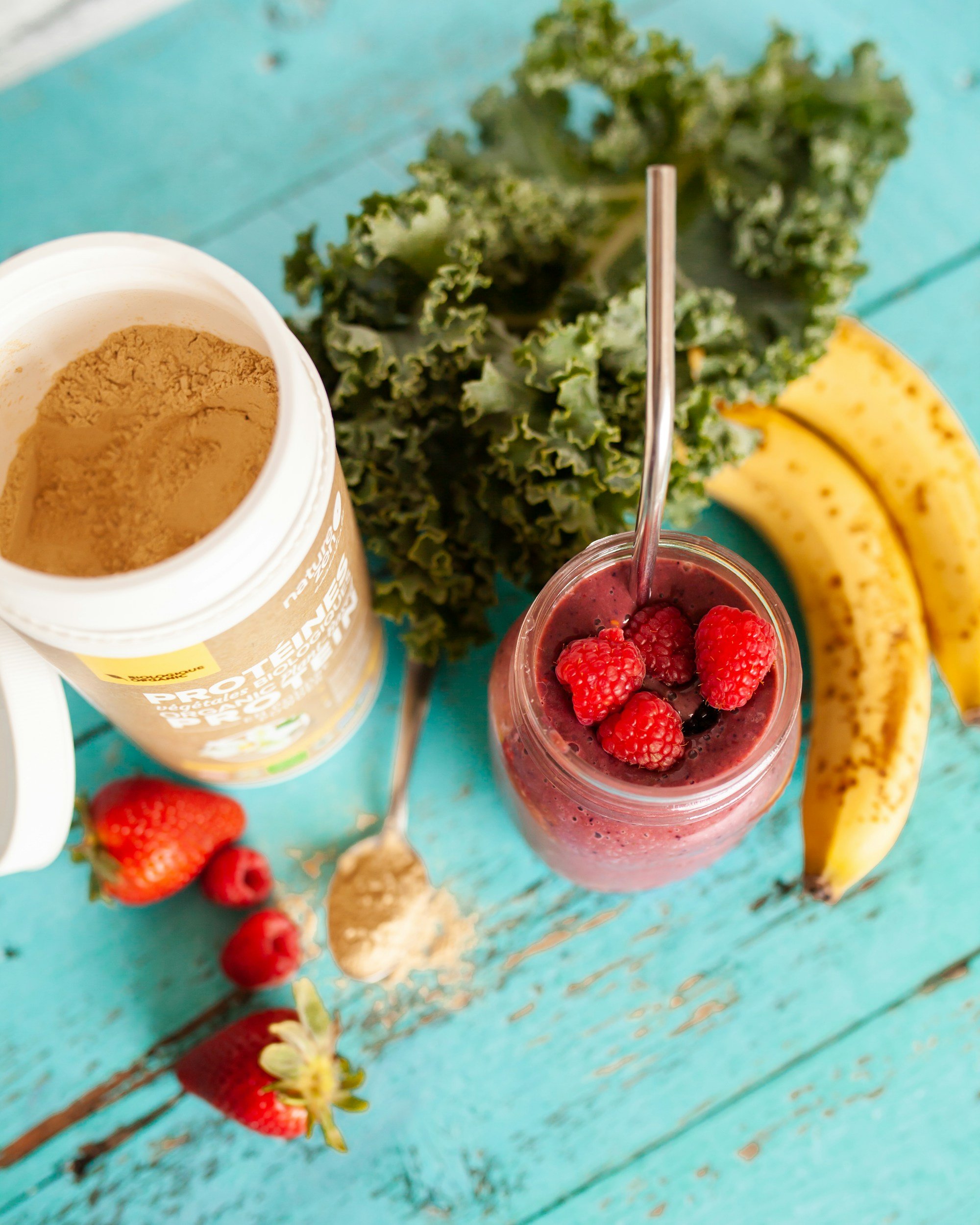 Protein powder, berry smoothie jar, banana, kale, and strawberries on a teal wooden table