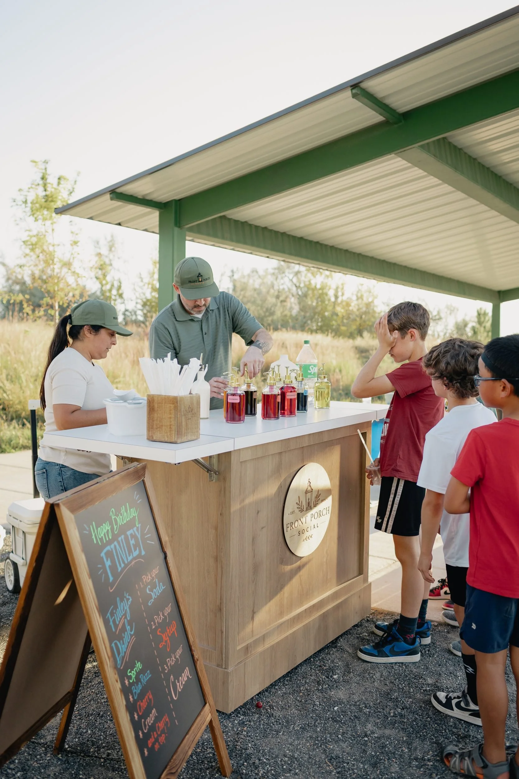 Two Front Porch Social servers making dirty sodas at the wooden soda cart under a park pavilion as kids line up at a birthday party