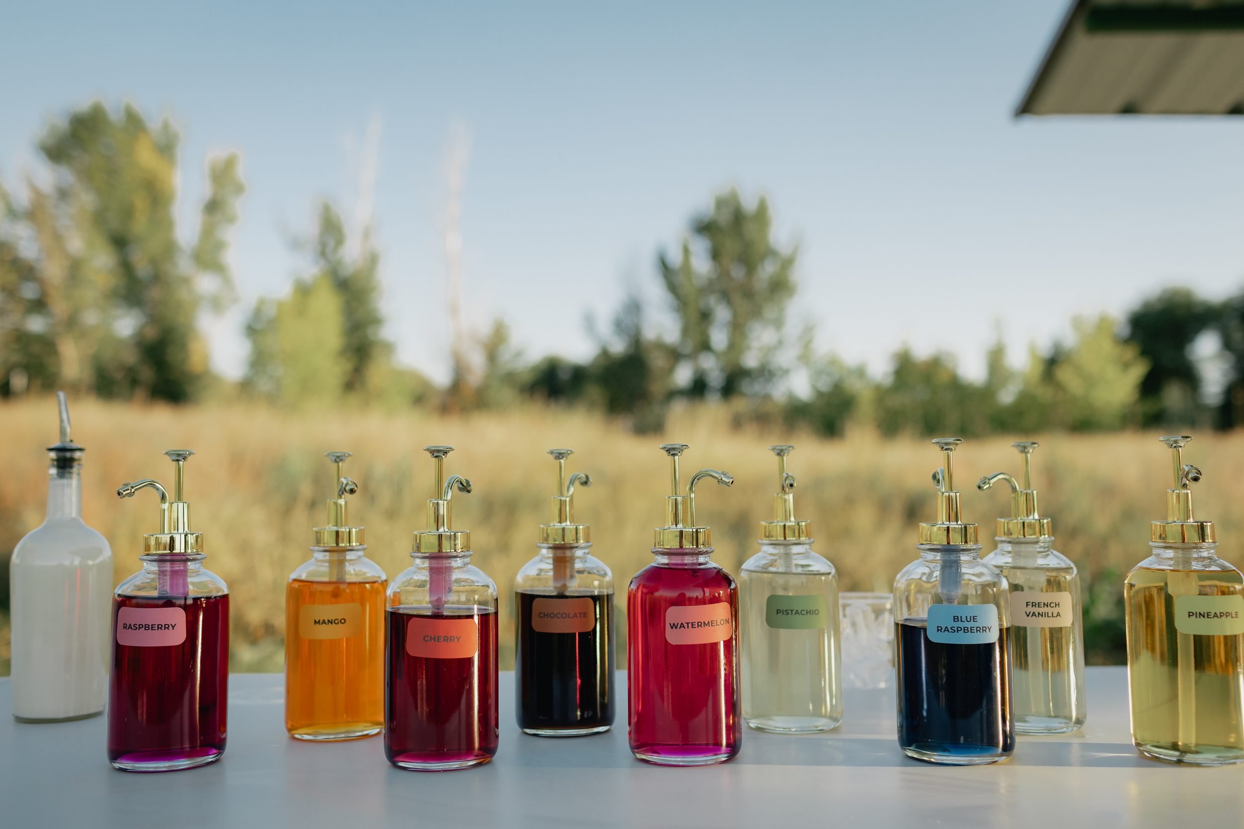 Row of labeled dirty soda syrups including raspberry, mango, cherry, chocolate, watermelon, pistachio, blue raspberry, French vanilla and pineapple, with a bottle of cream, outdoors