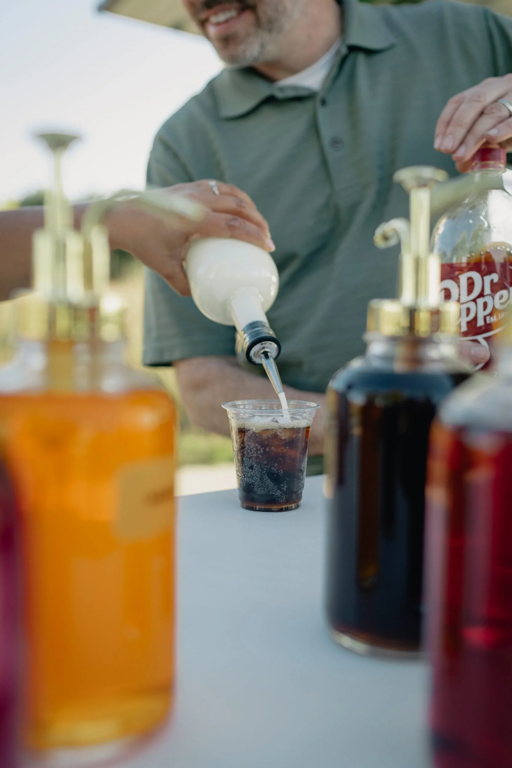 Cream being poured into a cup of Dr Pepper over ice to make a dirty soda, framed by colorful syrup bottles