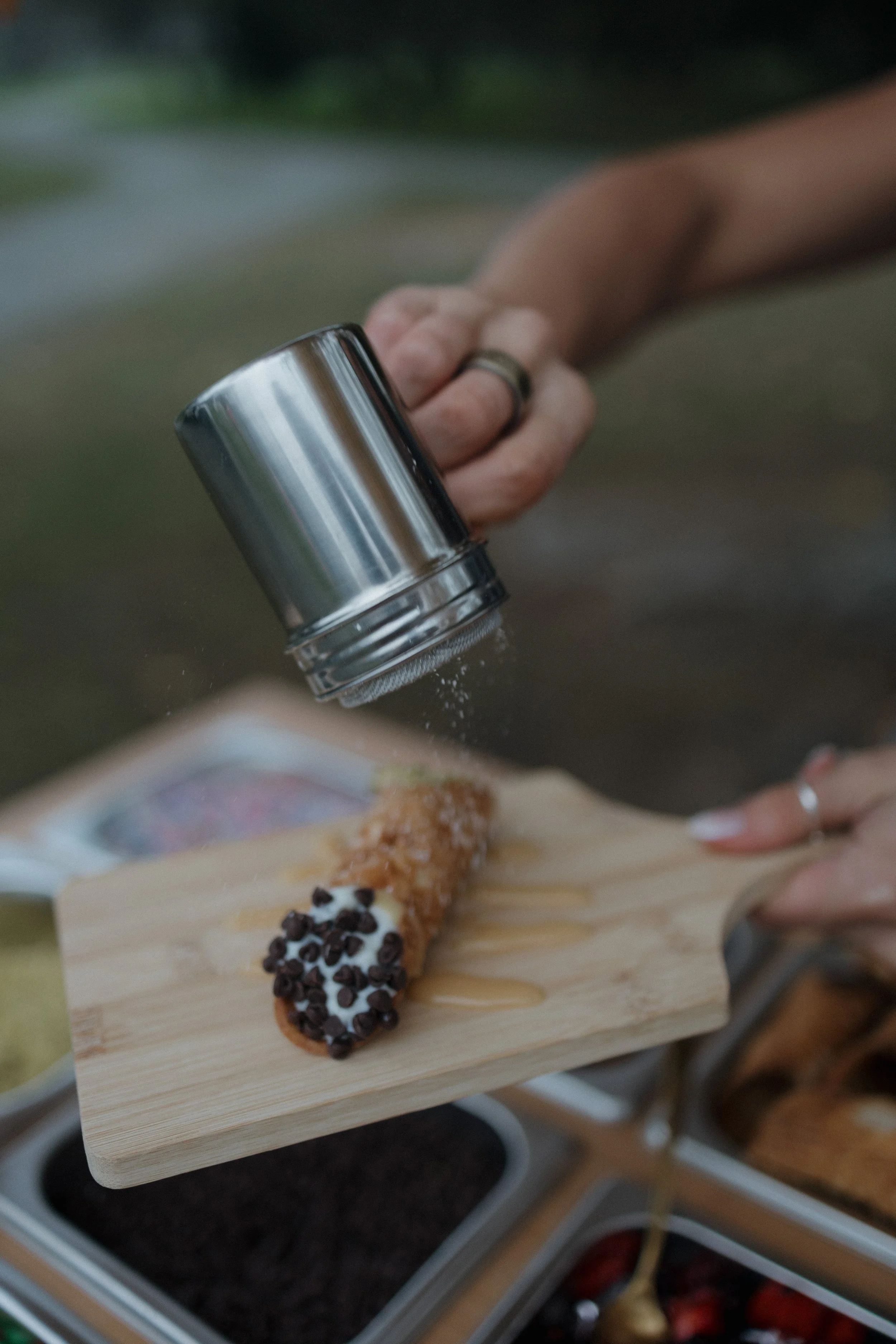 Cannoli on a wooden board being sprinkled with powdered sugar with wells of toppings in the background