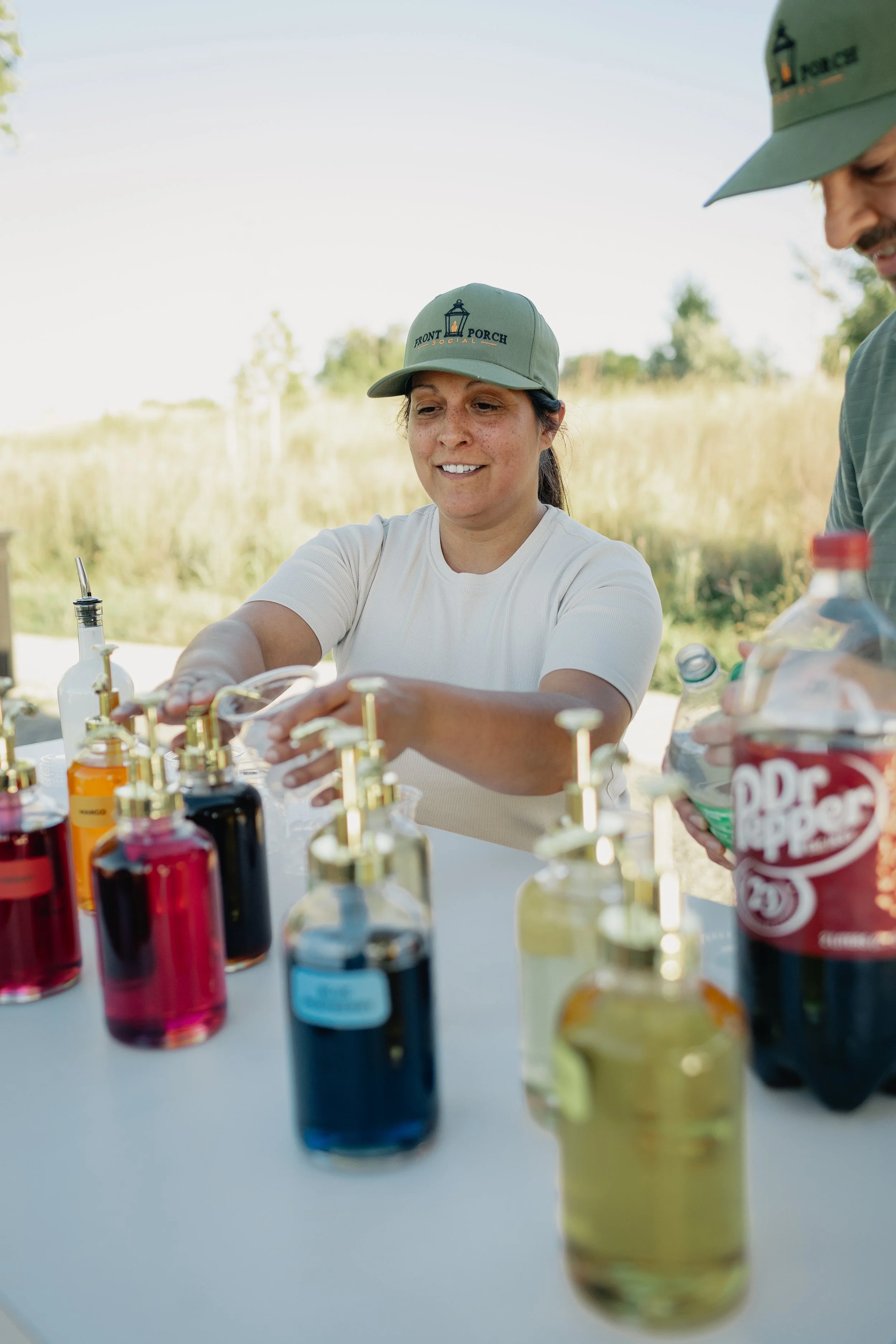 Smiling Front Porch Social server in a branded green cap building a dirty soda among gold-pump syrup bottles and a bottle of Dr Pepper