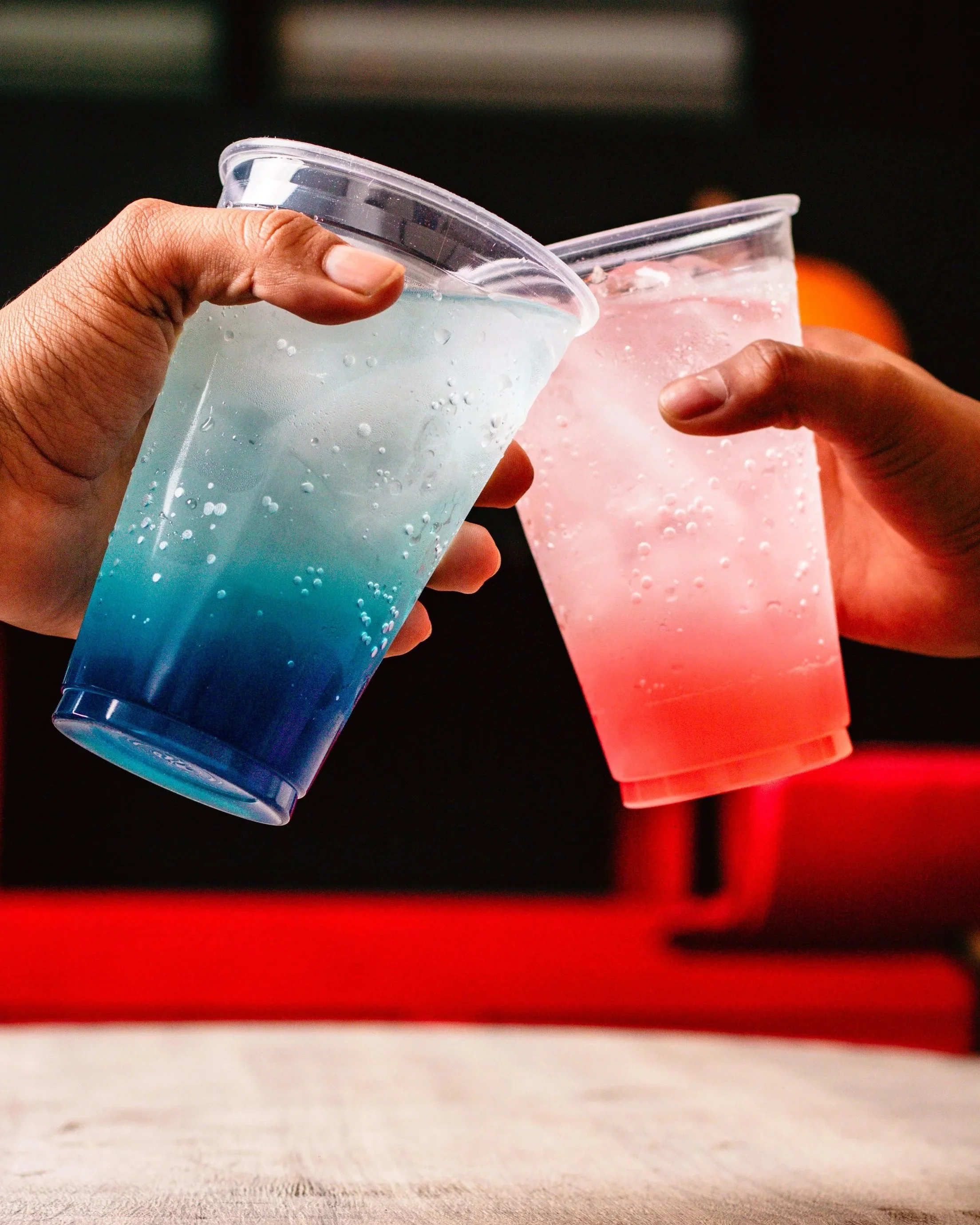 Two hands clinking colorful dirty soda drinks in plastic cups