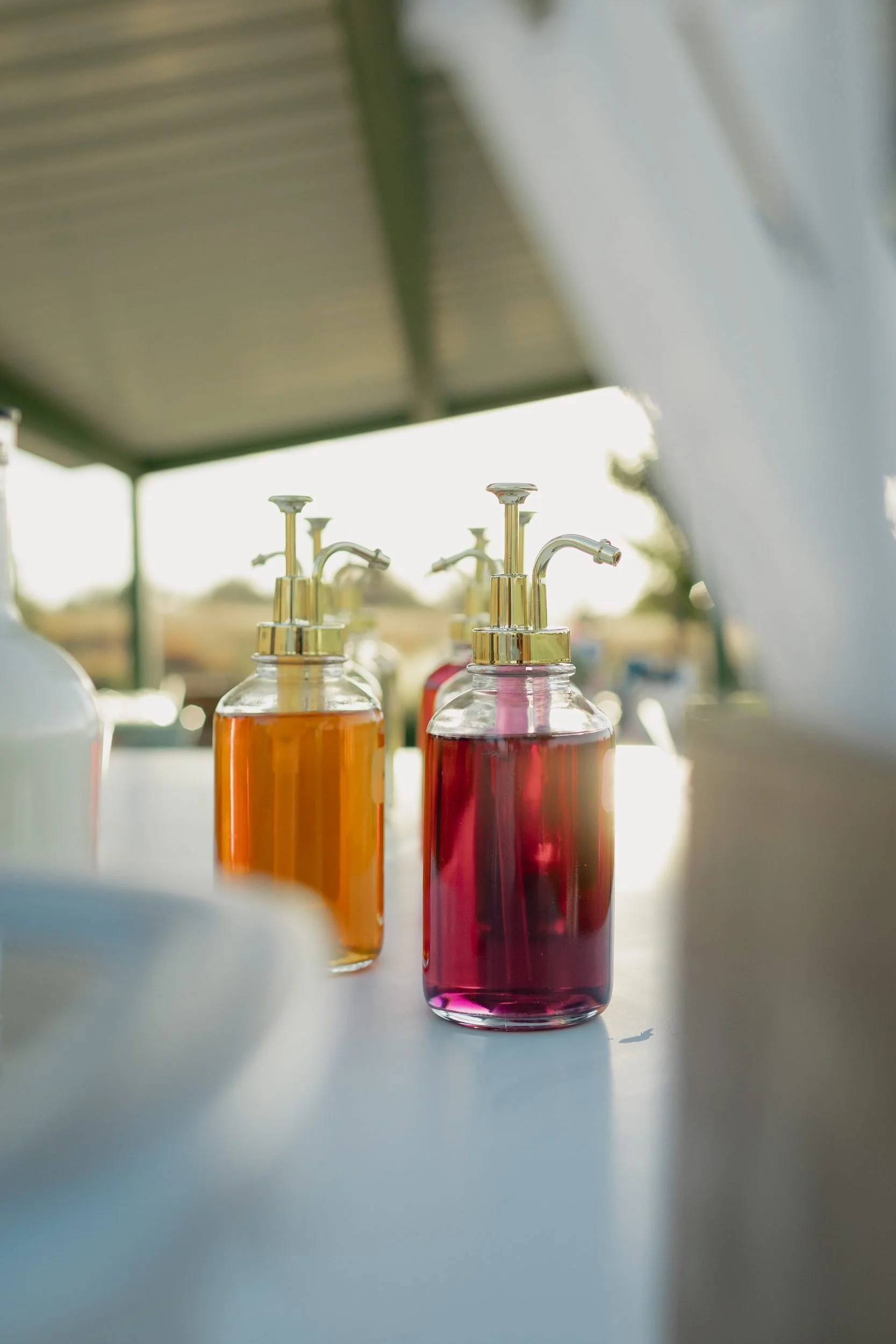 Mango and raspberry syrup bottles with gold pumps glowing in evening sunlight on the Dirty Soda Bar counter