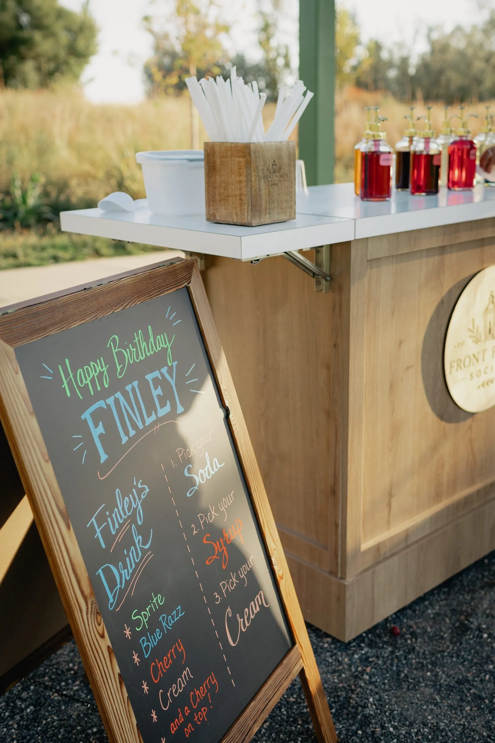 Hand-lettered chalkboard sign reading Happy Birthday Finley with a pick-your-soda, syrup and cream menu beside the wooden Front Porch Social soda cart