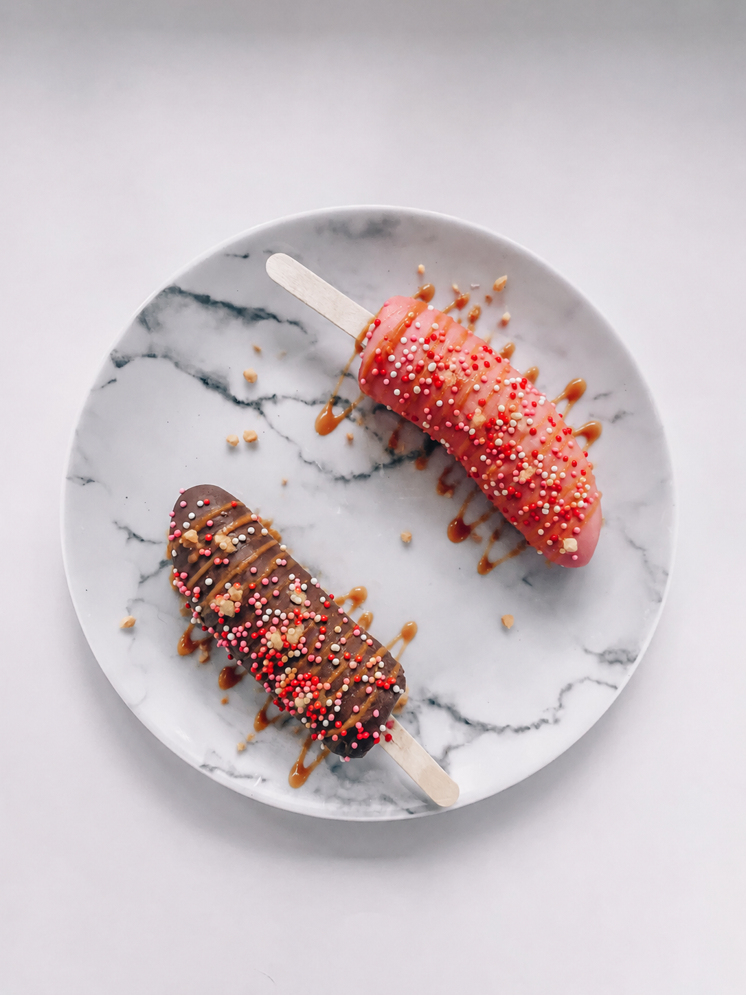 Two chocolate-dipped frozen banana pops with colorful toppings on a marble plate