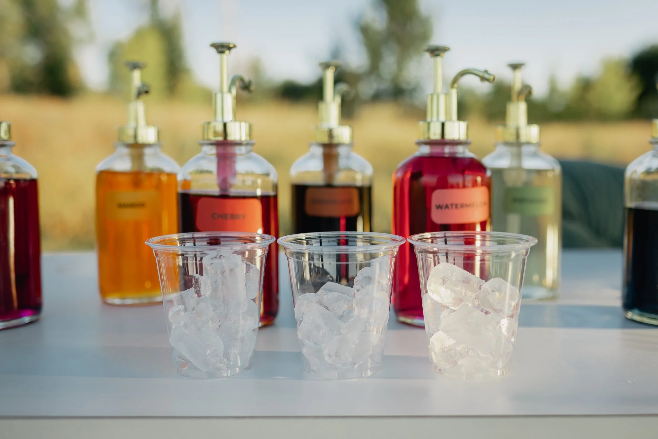 Three clear cups of ice in front of mango, cherry, chocolate, watermelon and pistachio syrup bottles at the Dirty Soda Bar