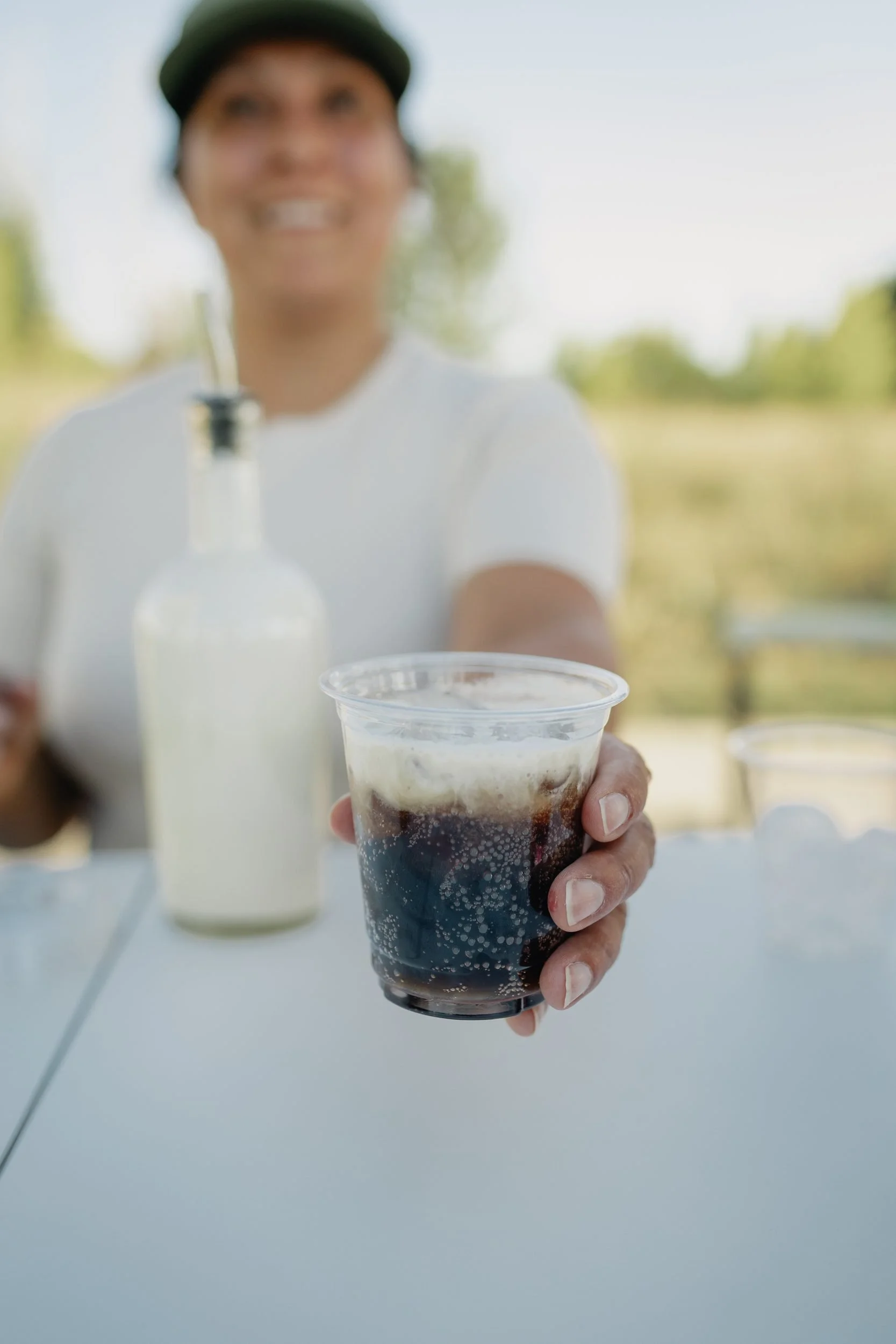 Front Porch Social server handing over a dirty soda in a clear cup, layered with cream over dark cola, at the mobile Dirty Soda Bar in Northern Colorado