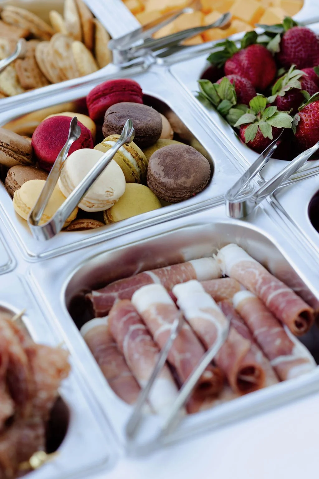 Meat, fruit and desserts in small wells with tongs on a mobile charcuterie cart with a white top.
