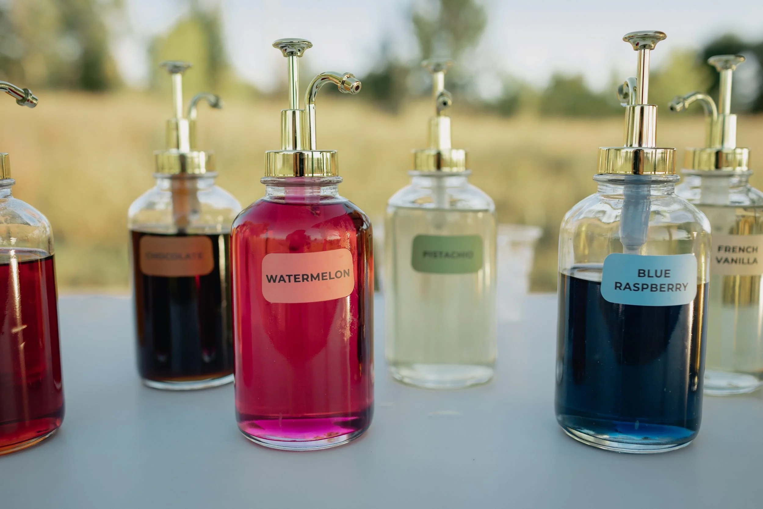 Close-up of labeled watermelon, chocolate, pistachio, blue raspberry and French vanilla syrup bottles with gold pumps on the Dirty Soda Bar