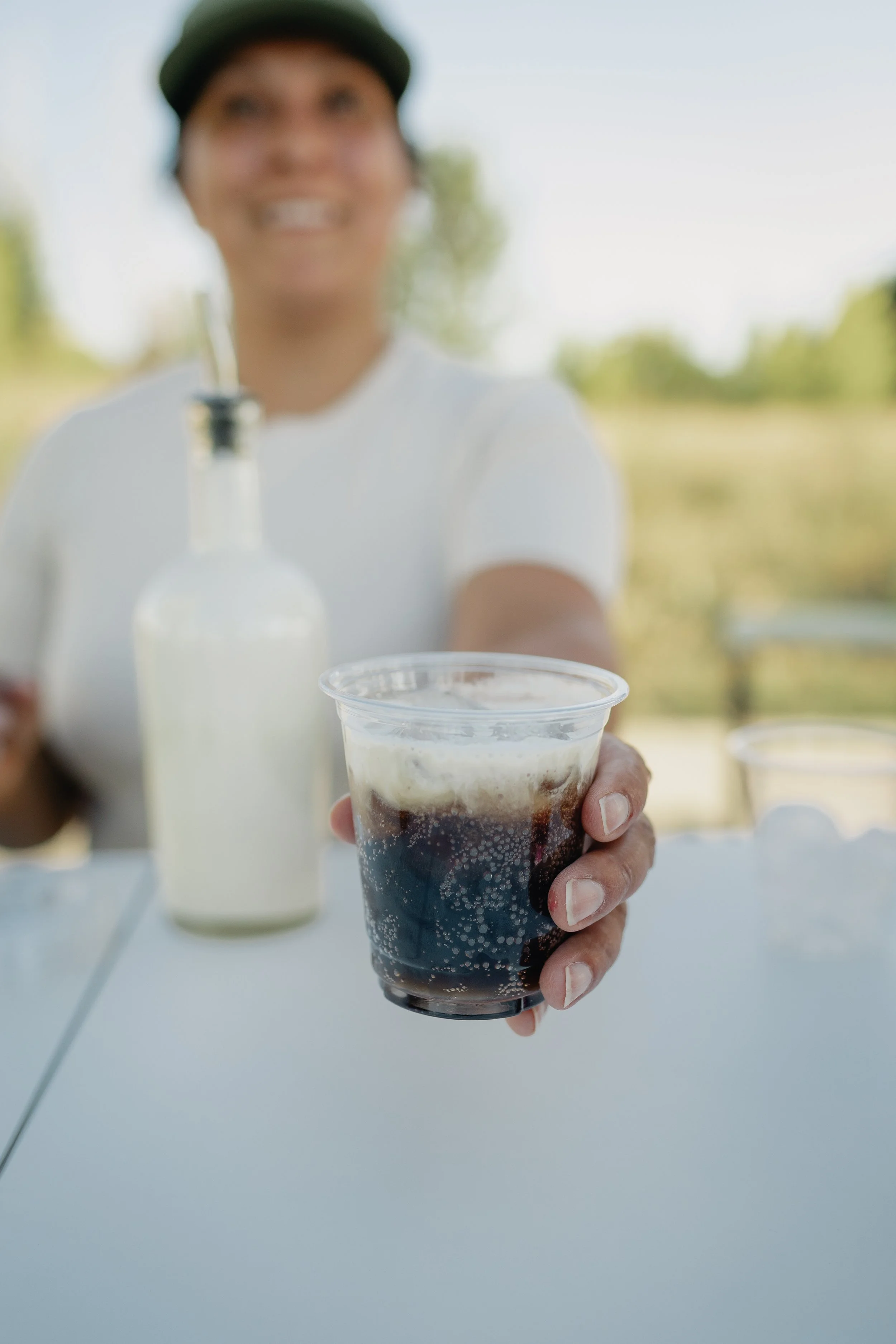 Front Porch Social server handing over a dirty soda in a clear cup, layered with cream over dark cola, at the mobile Dirty Soda Bar in Northern Colorado