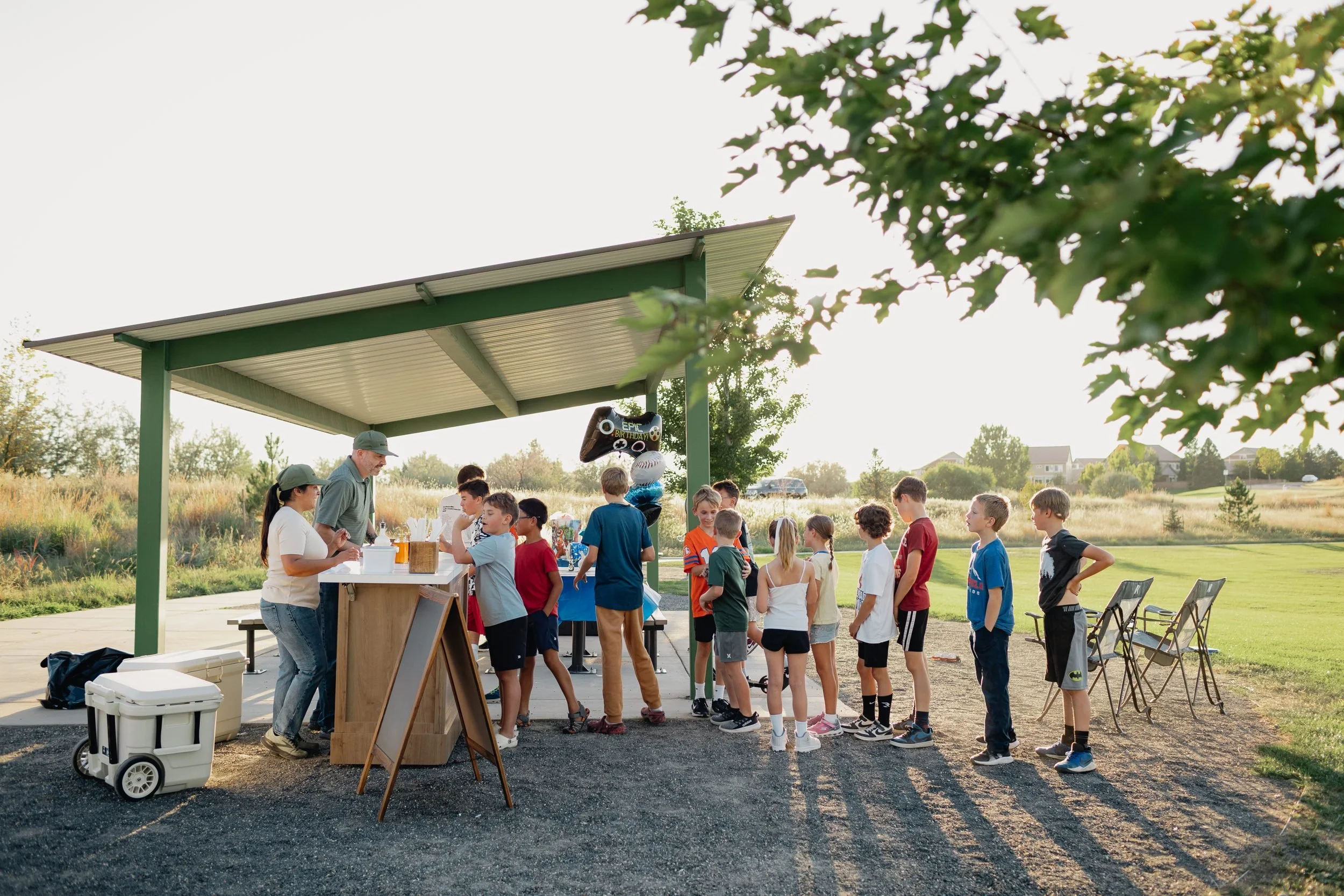 Long line of kids waiting at the Front Porch Social Dirty Soda Bar under a green park pavilion during a birthday party in Northern Colorado