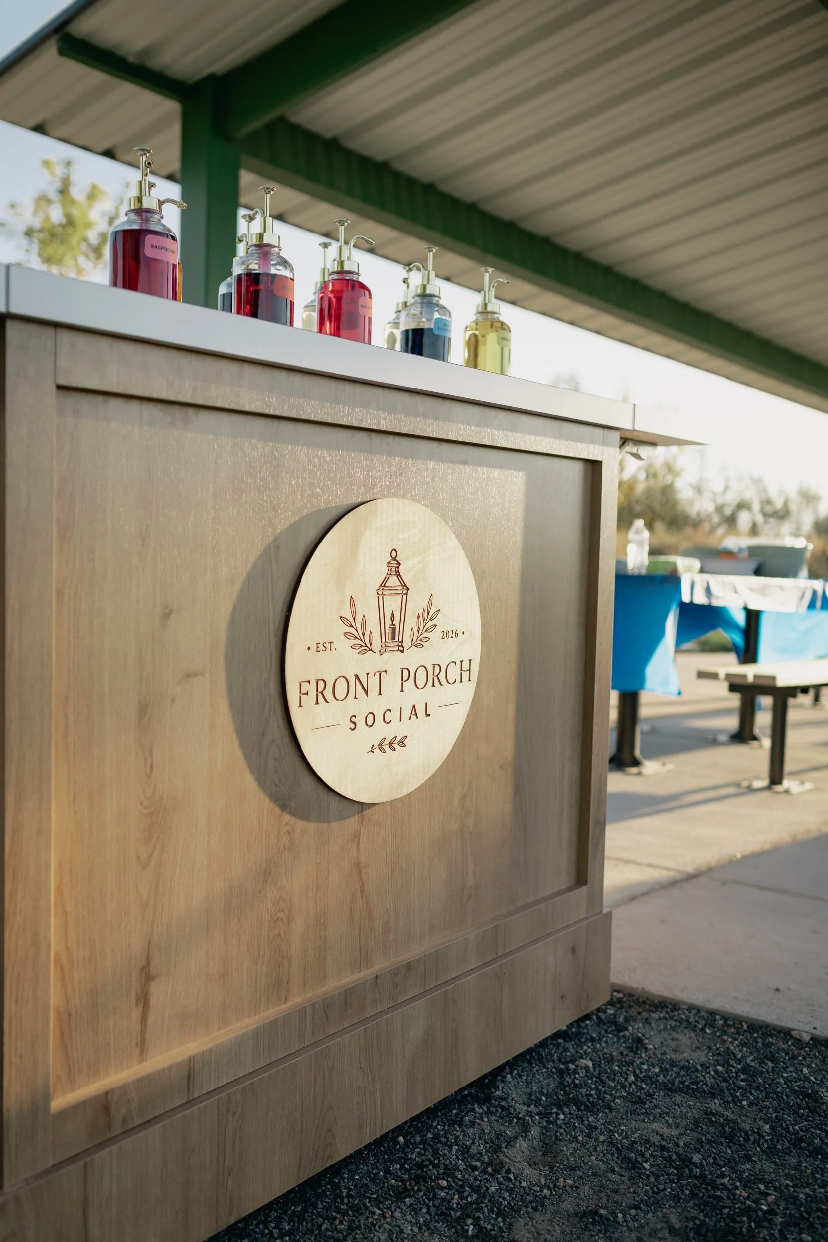 Wooden Front Porch Social mobile soda cart with an engraved round logo sign and colorful syrup bottles on top, set up under a park pavilion