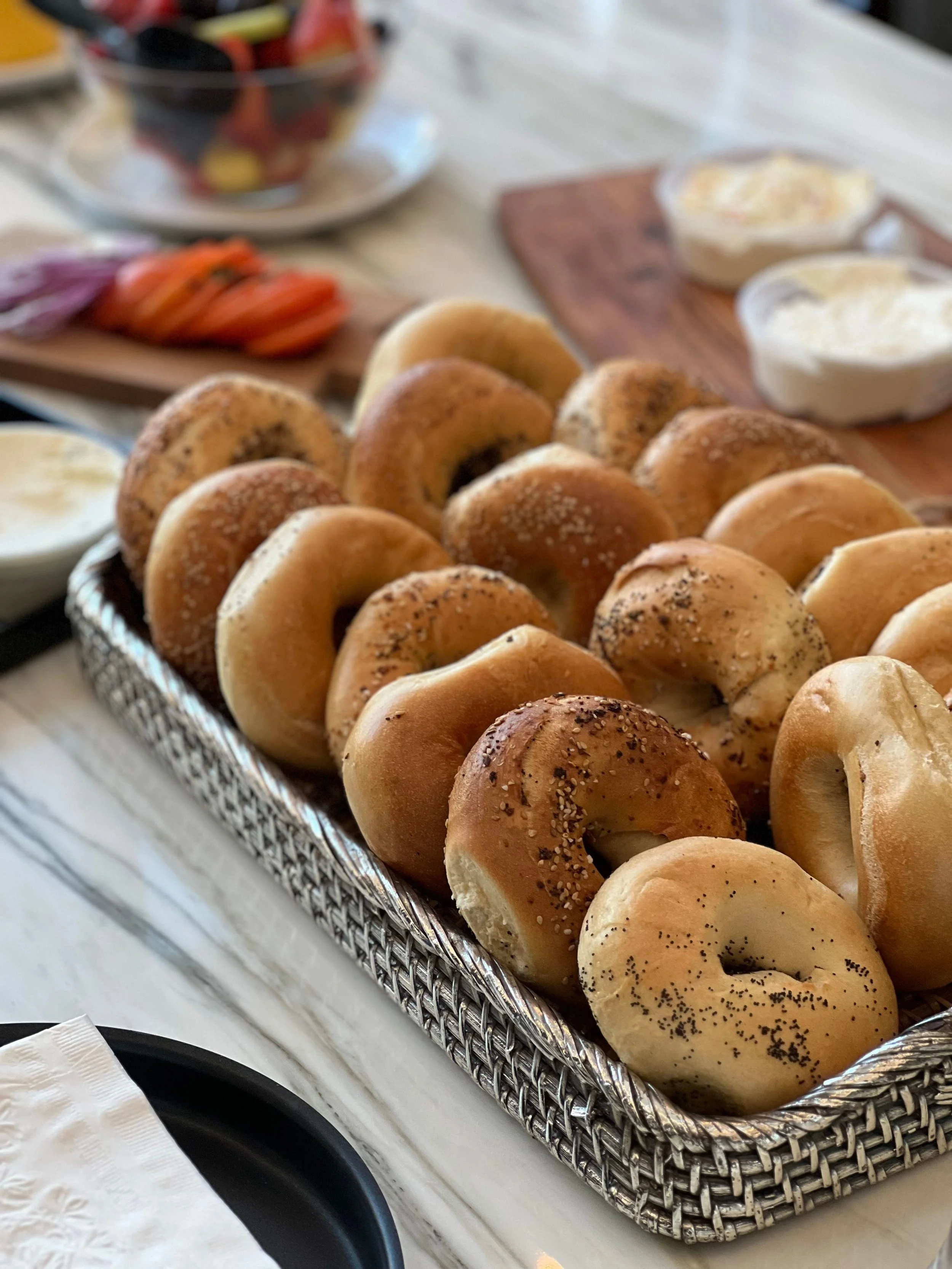 Assorted bagels arranged in a woven basket alongside sliced vegetables and spreads