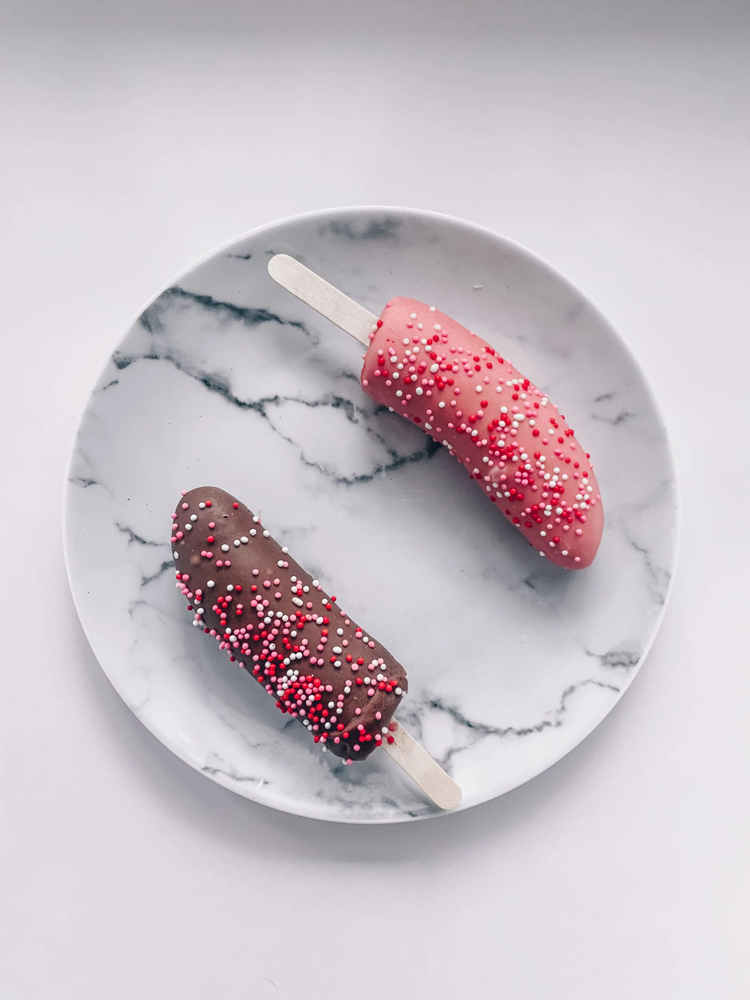 Two chocolate and berry-coated frozen banana pops on a marble plate