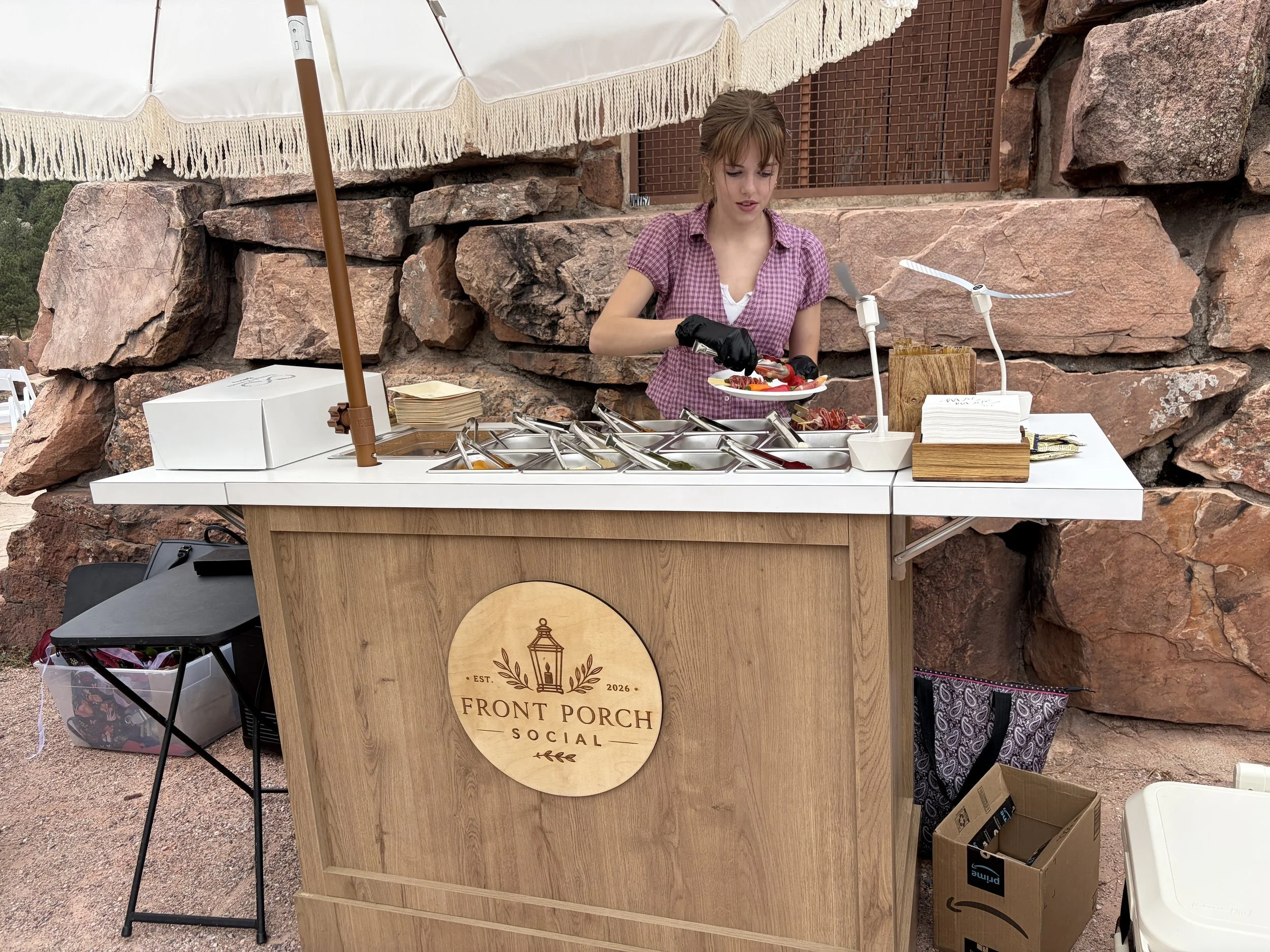 Front Porch Social charcuterie cart with fringed umbrella serving guests at an outdoor Colorado wedding among red rocks