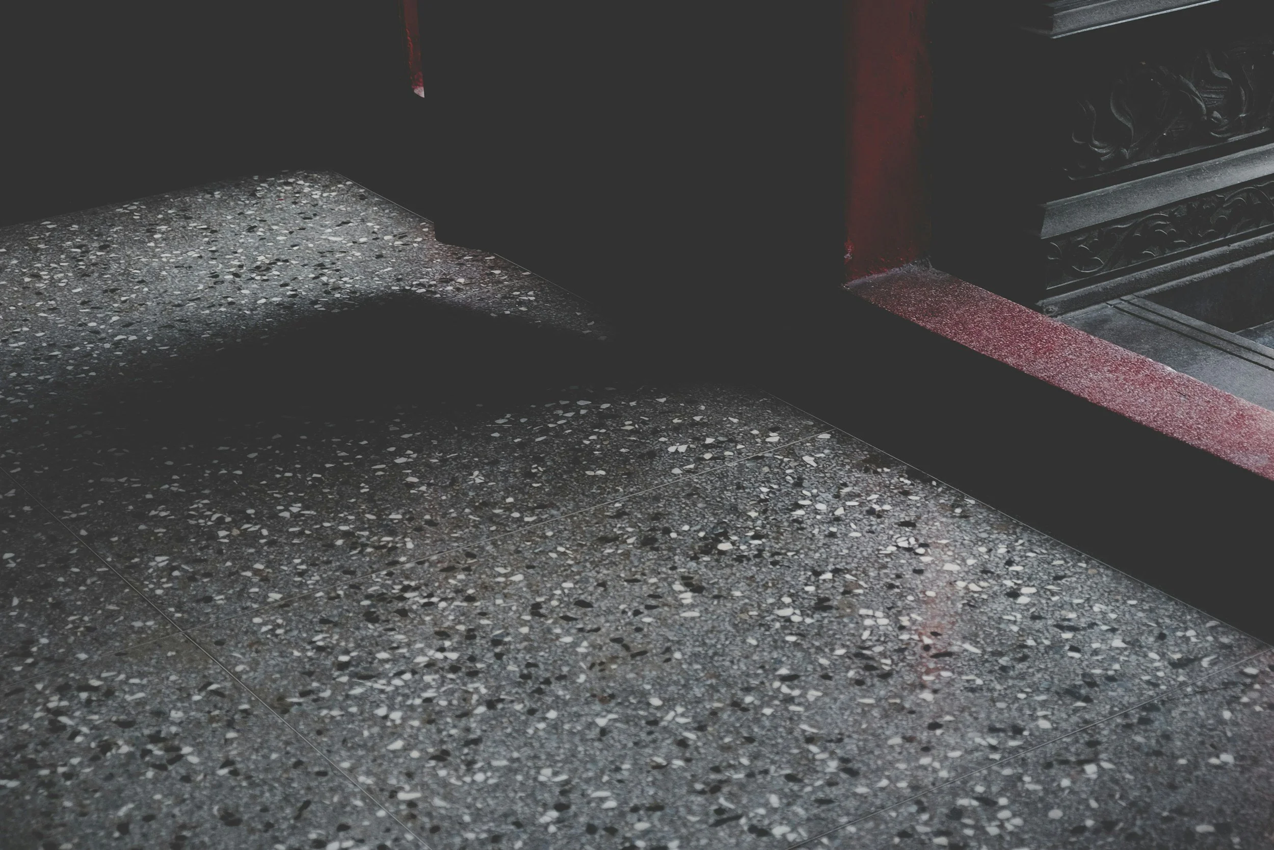 Close-up of a textured gray terrazzo floor with a black door and a red-painted step or curb in the background.