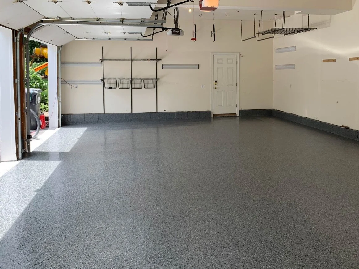 Empty garage with open door, clean gray epoxy floor, white walls, shelving and hooks mounted on the back wall, and a closed door to the interior.