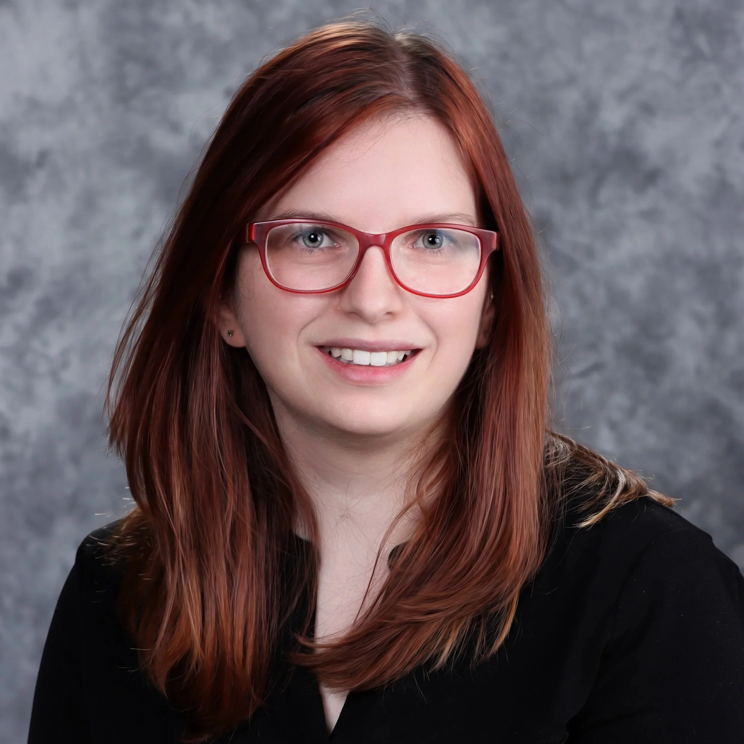 Amy Chodorowski Pmhnp Illinois Close-up of a woman with red hair wearing red glasses, smiling, against a gray textured background.