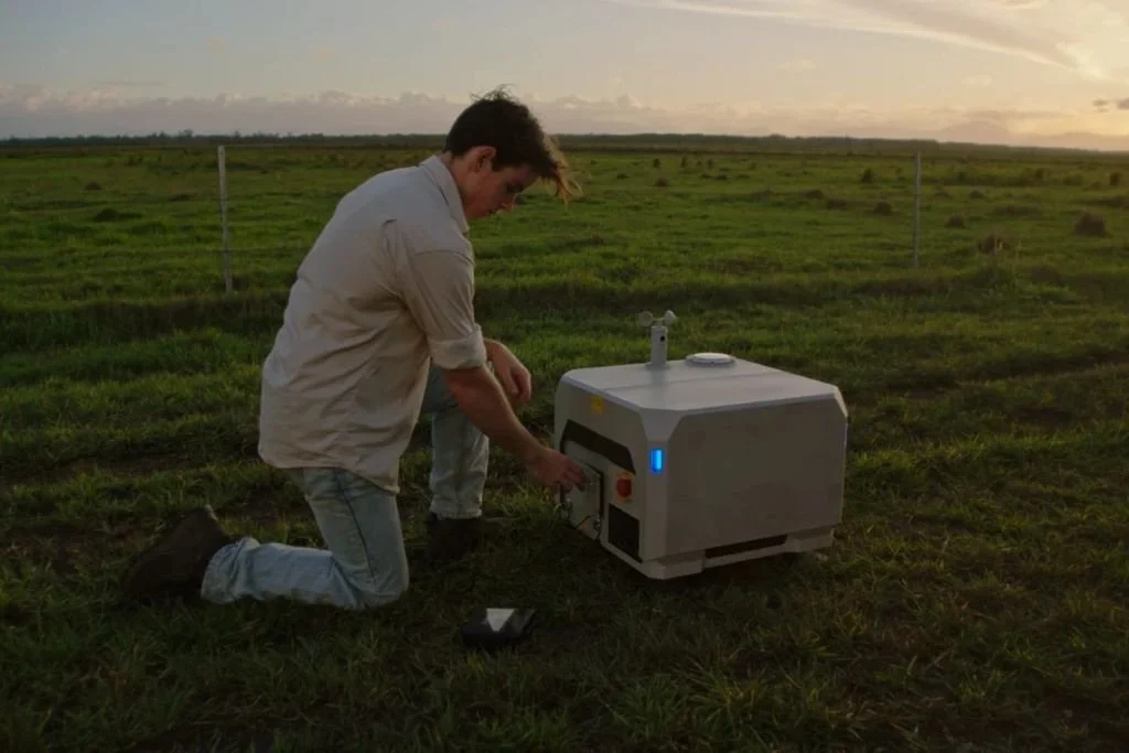 Image of Sam Rogers Brumby, A man kneeling on grass in an open field at sunset, working on a piece of equipment, who was helped by Beanstalk AgTech and Monsoon Ventures