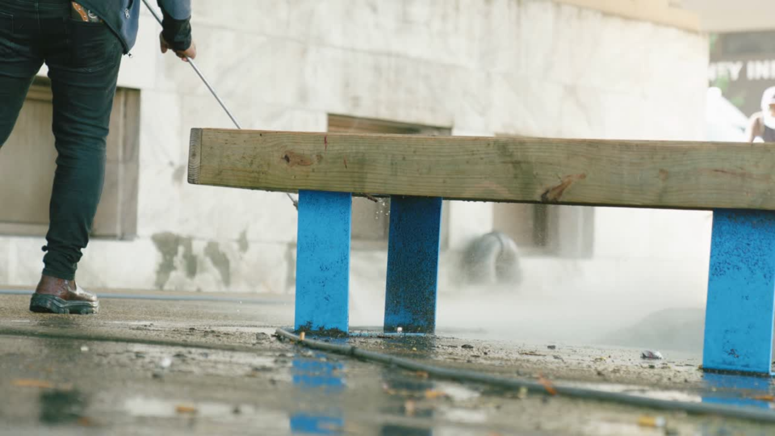 Person in black pants and shoes using a crowbar to remove a wooden bench painted blue, on a sidewalk near a building.