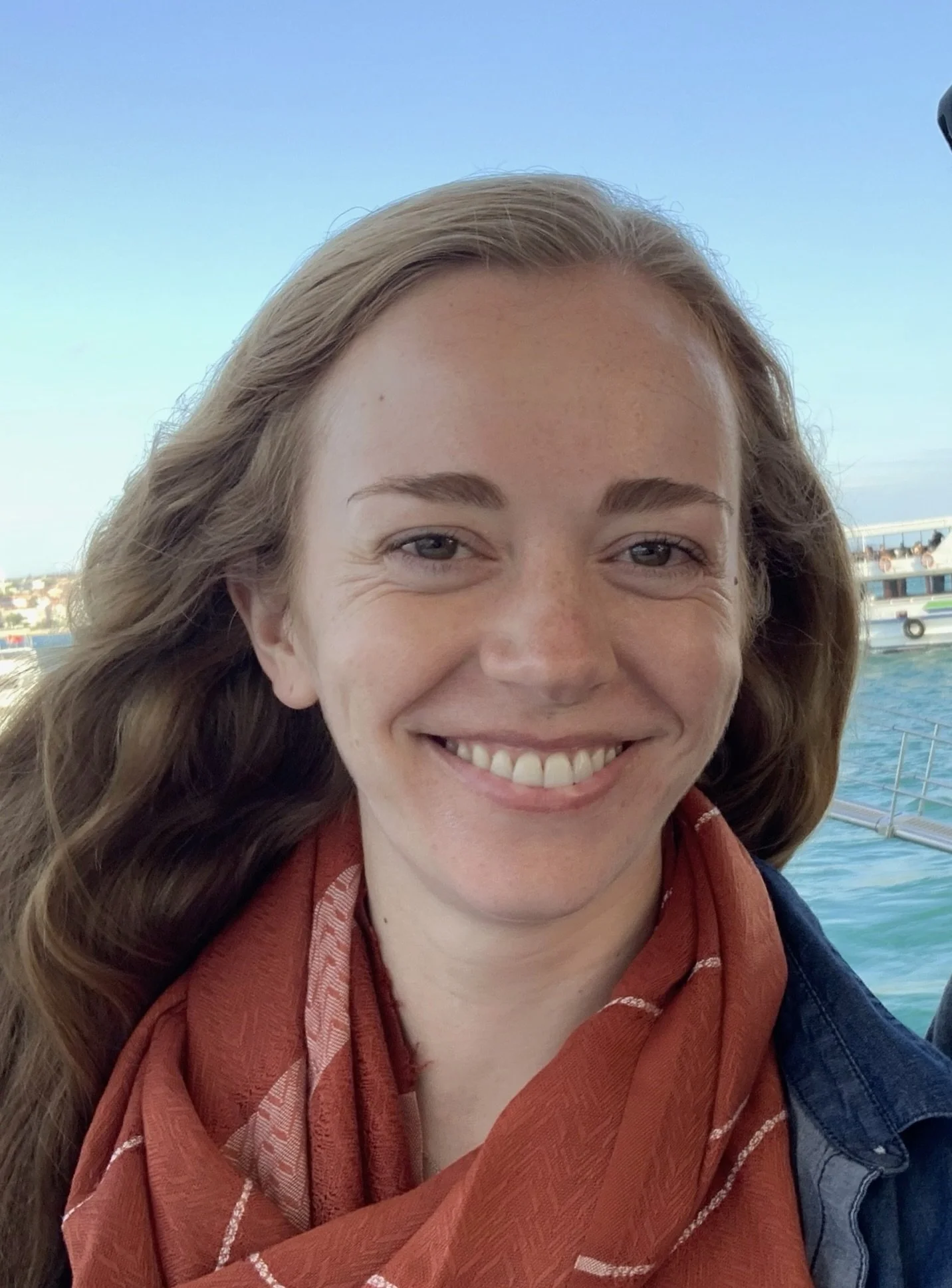 Close-up of a smiling young woman with long, wavy hair, standing outdoors near water, wearing a red scarf and a jacket, with a boat and cityscape in the background.