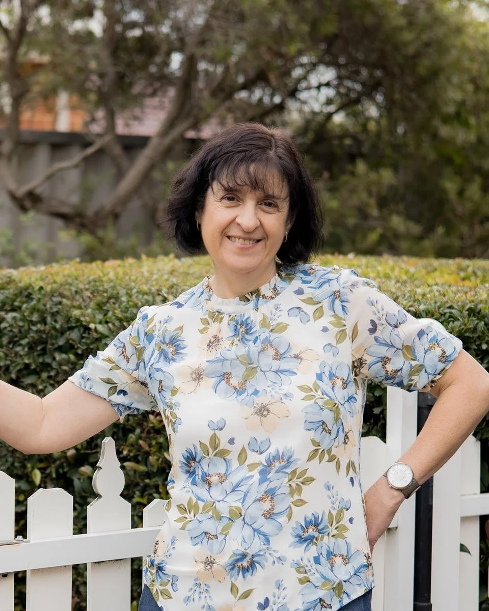 Portrait of Mechelle Palermo in a floral blouse smiling outdoors next to a white picket fence with trees in the background.