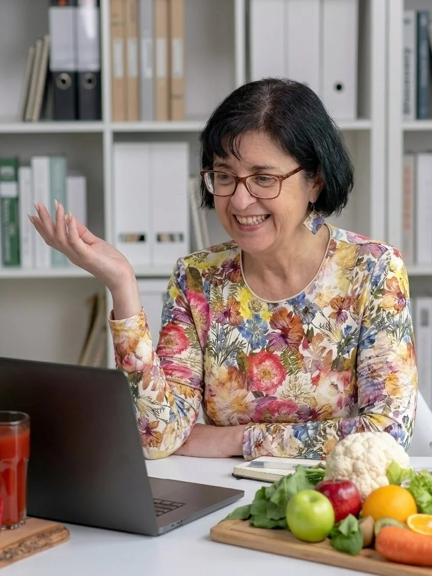 A woman with glasses smiling and gesturing while sitting at a table with a laptop, a notebook, a glass of red juice, and a wooden cutting board with fruits and vegetables, including cauliflower, apples, oranges, and lettuce, in an office setting.