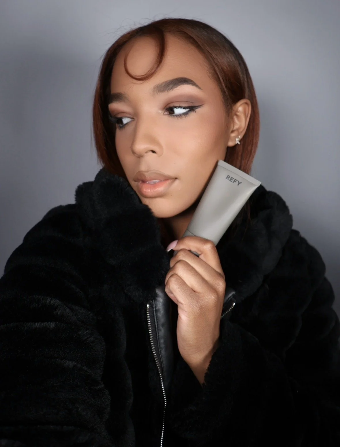 A young woman with short brown hair, makeup, and earrings holding a gray tube labeled 'REFY' close to her face, posing in a black fur jacket against a plain background.