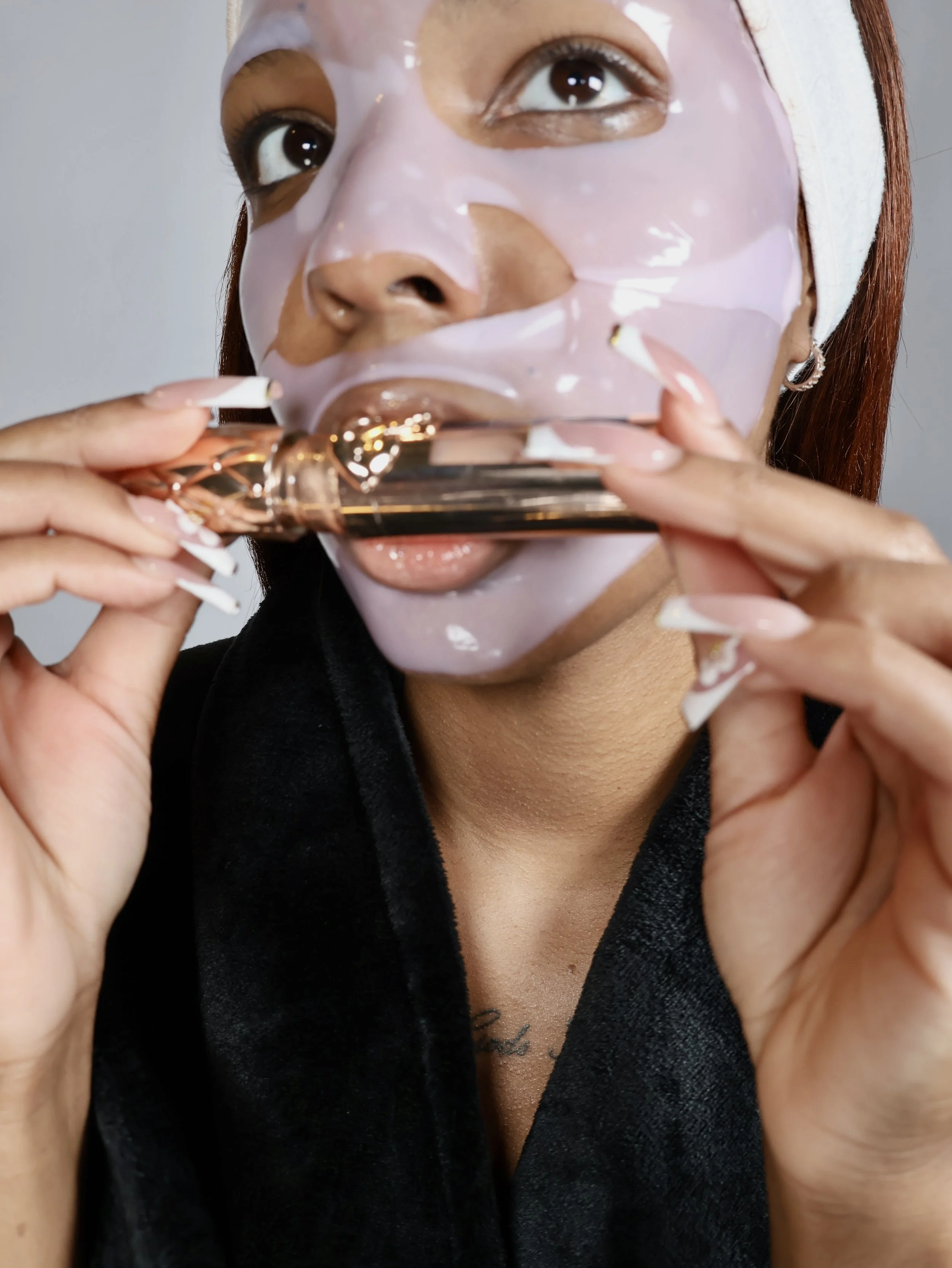 A woman with reddish hair, wearing a white headband, is peeling off a pink facial sheet mask with gold accents from her face.