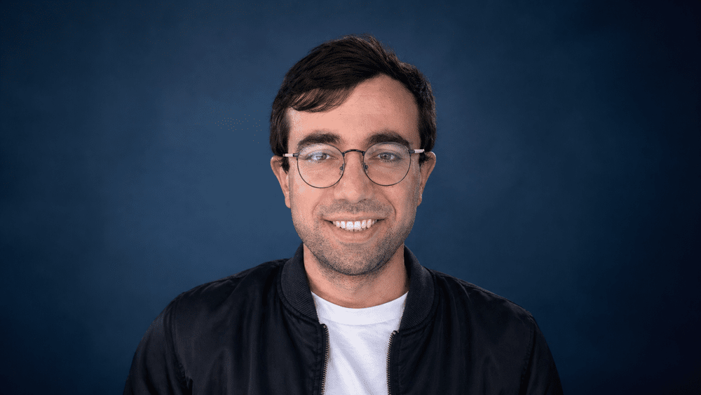 Portret van een glimlachende man met bruin haar, bril, zwarte jas en witte T-shirt, voor een donkere blauwe achtergrond.