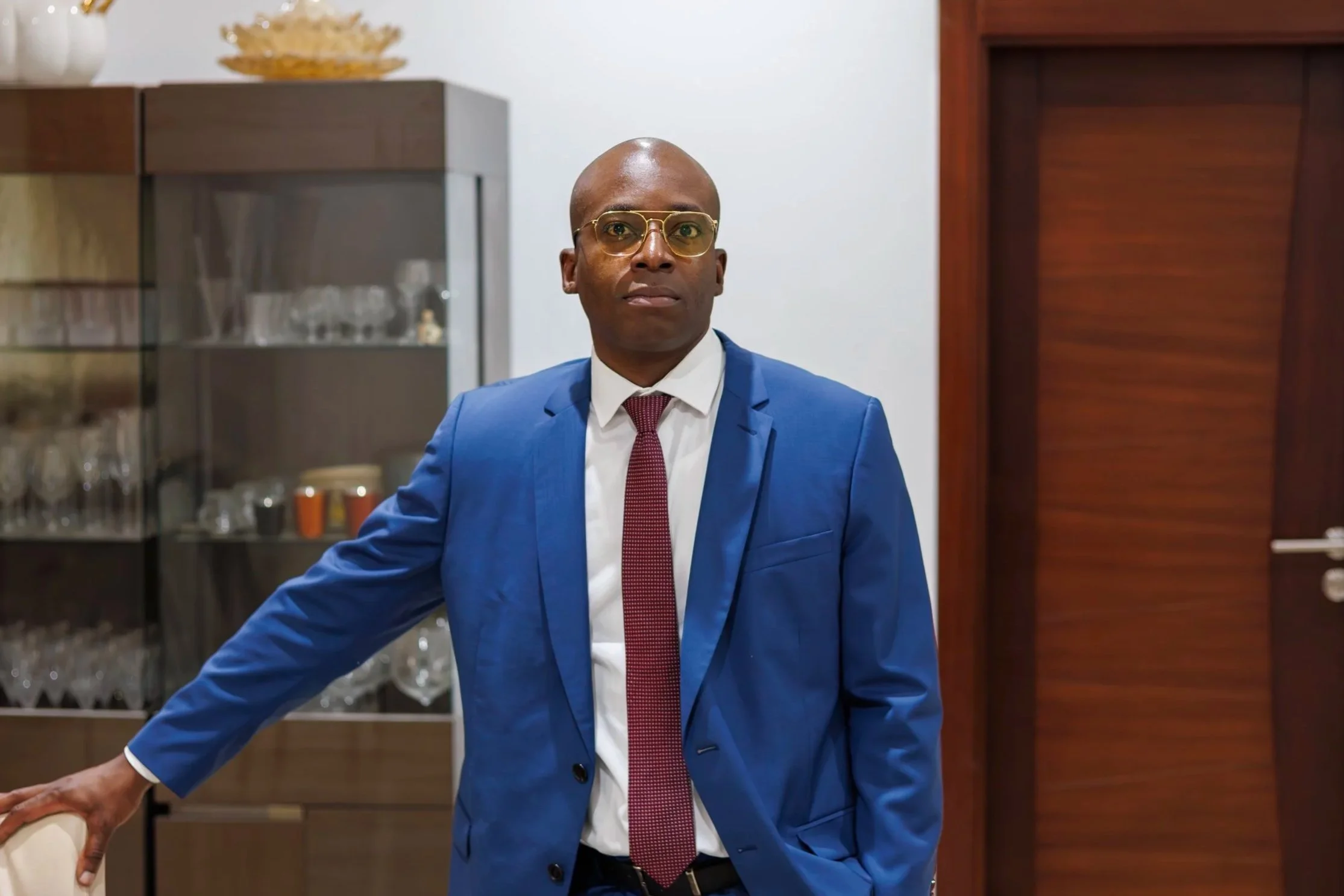 A man in a blue suit with gold-rimmed glasses, standing in a modern room with a glass-fronted cabinet and wooden shelves behind him.