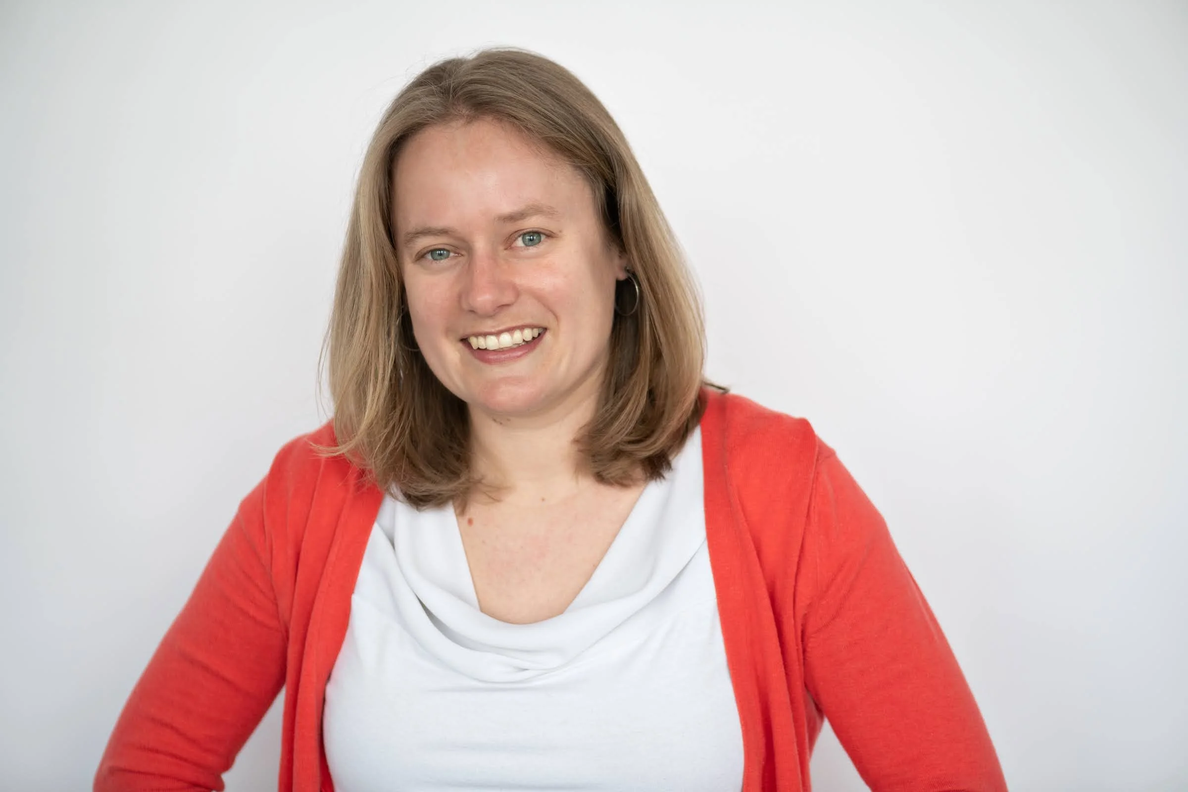 Emma Bohmann, a woman with shoulder-length brown hair smiling, wearing a white shirt and red cardigan against a plain white background.