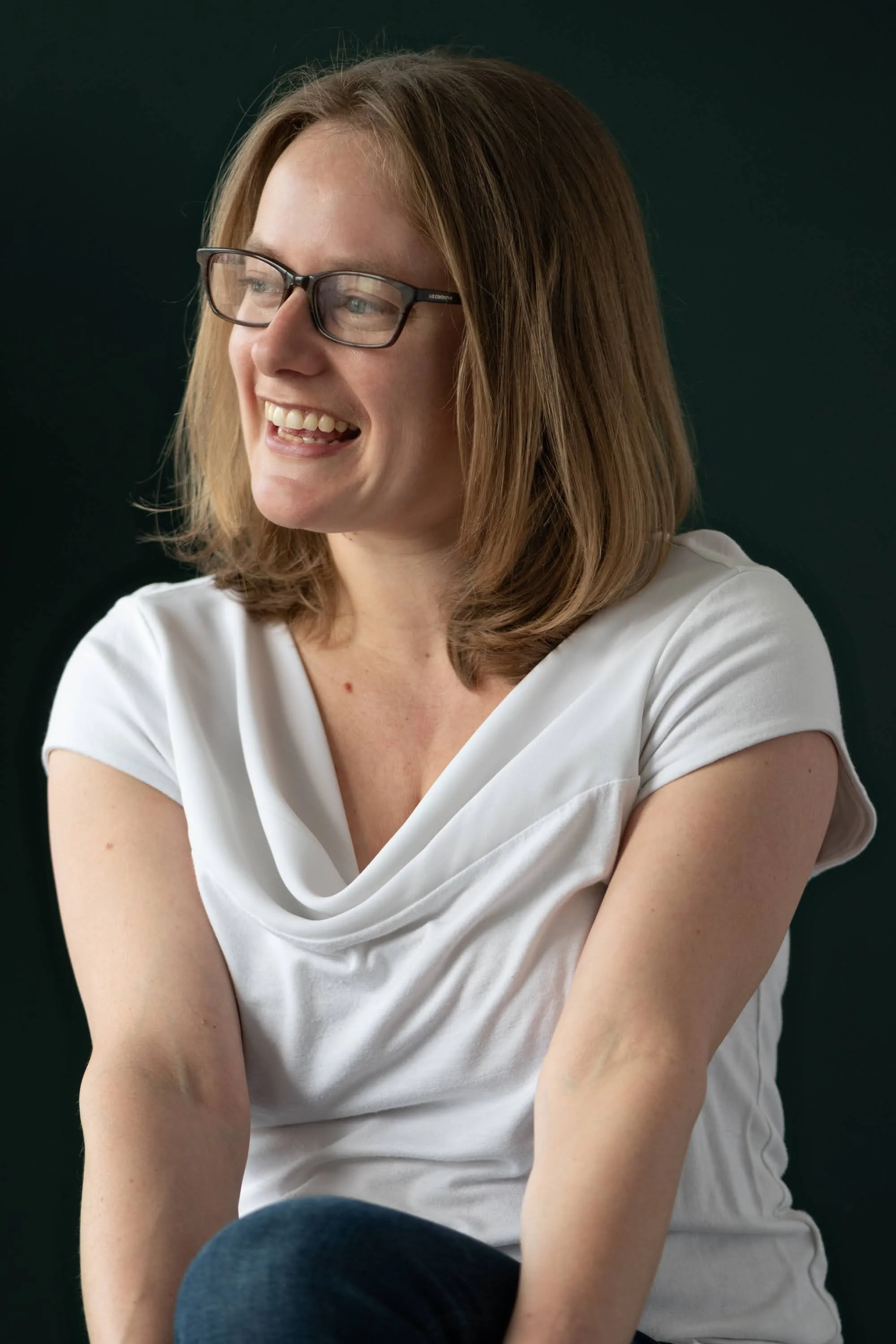 Emma Bohmann, a woman with shoulder-length reddish-brown hair, wearing glasses and a white top, smiling and looking to her left. She is sitting with her arms crossed and appears to be in a happy or joyful mood. The background is dark green.