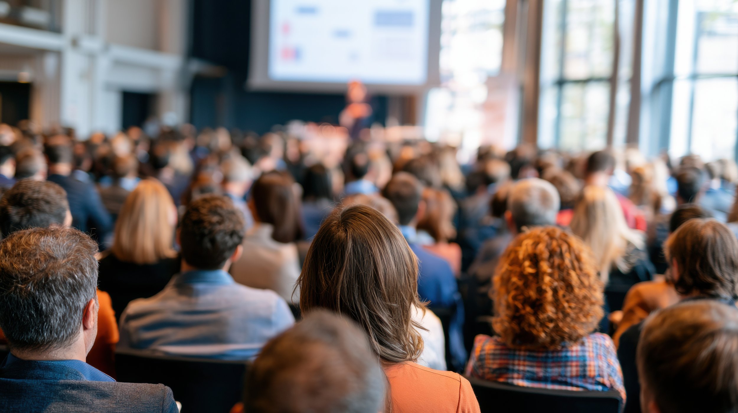 A large group of people attending a conference or seminar in a modern, well-lit room, with a presenter at the front near a large screen.