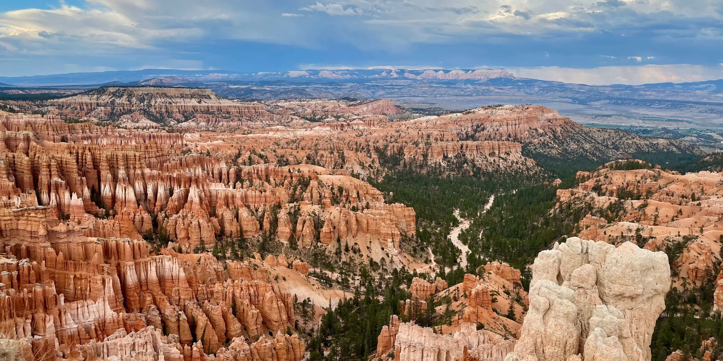A panoramic view of Bryce Canyon with orange and pink hoodoo rock formations, forests, and distant blue mountains under a partly cloudy sky.