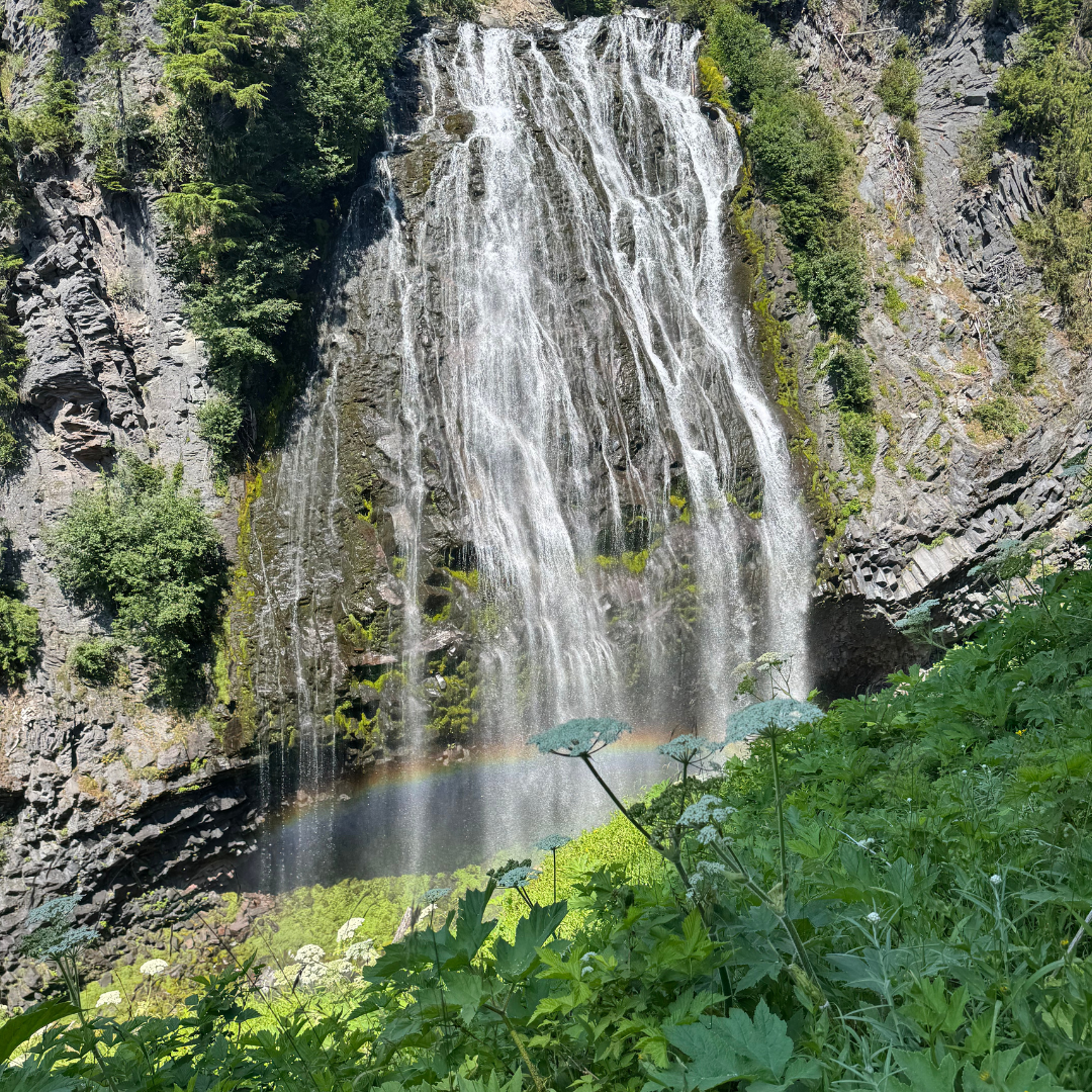 Waterfall cascading down a rocky cliff surrounded by green trees and foliage, with a faint rainbow visible at the base.