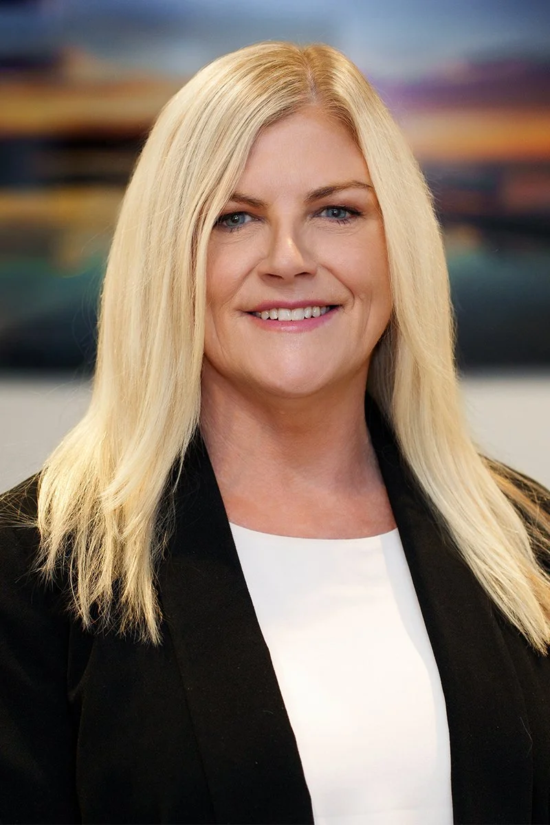 A blonde woman with straight hair, wearing a black blazer over a white top, smiling, standing indoors with a blurred abstract background.