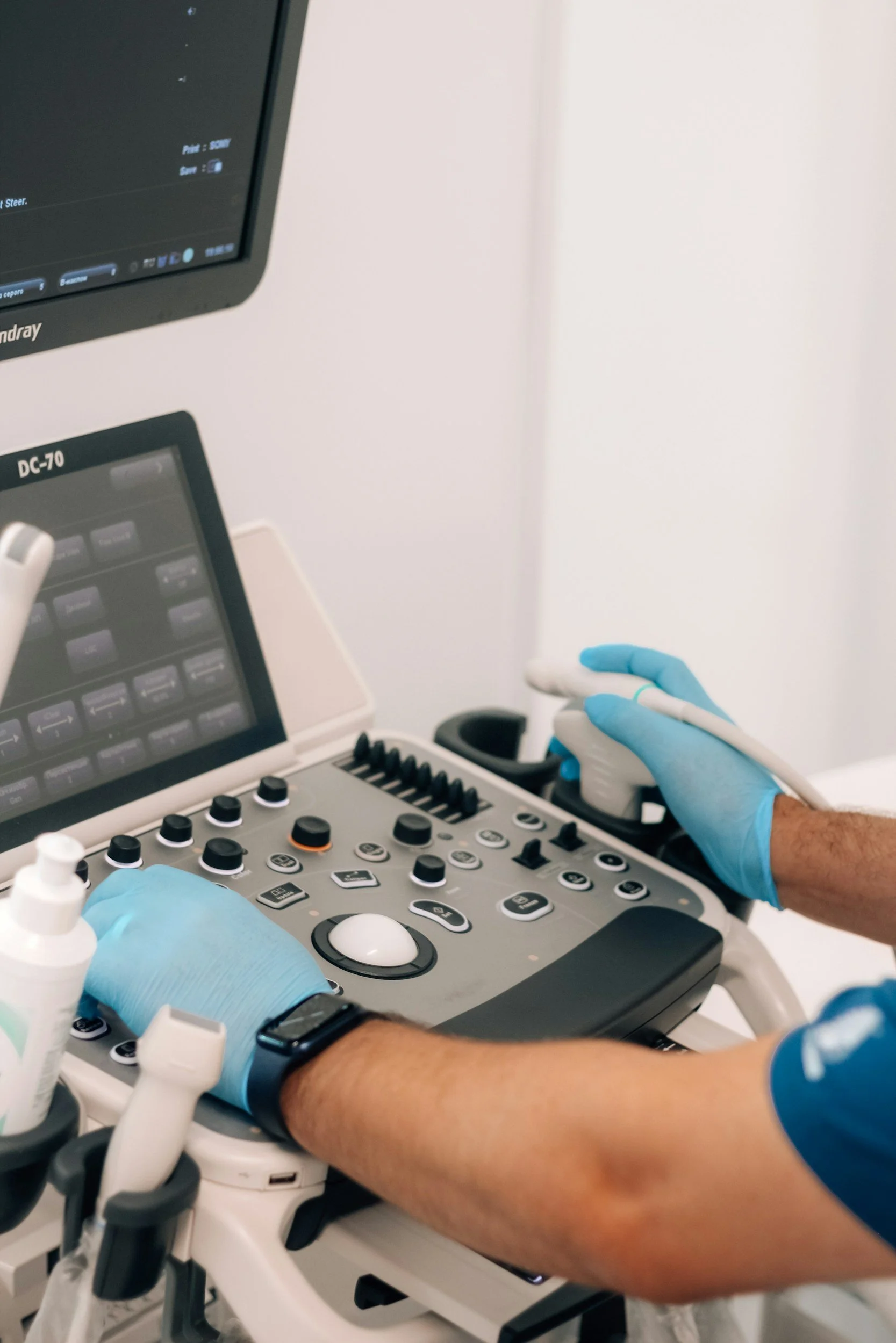 A medical professional in blue gloves operating an ultrasound machine in a clinical setting.