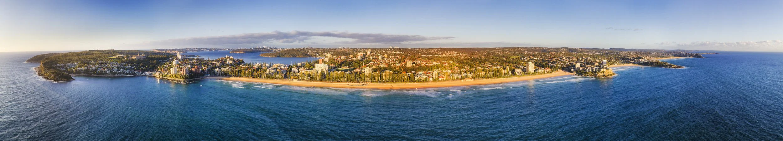 Aerial view of a coastal city with beaches, residential buildings, and greenery along the shoreline on a clear day.