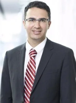 A man in a dark suit, white shirt, and red striped tie, wearing glasses, smiling in an office setting.