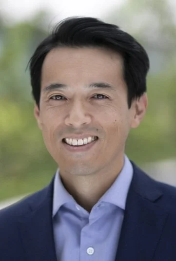 Headshot of a smiling Asian man in a navy blazer and light blue shirt outside with blurred greenery in the background.