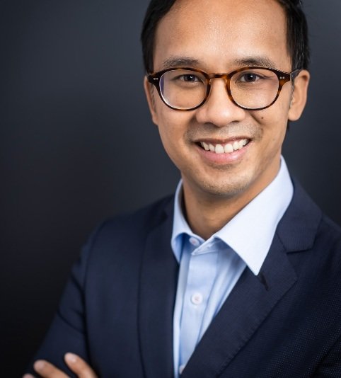 A professional man with glasses, wearing a navy blazer and light blue shirt, smiling against a dark background.