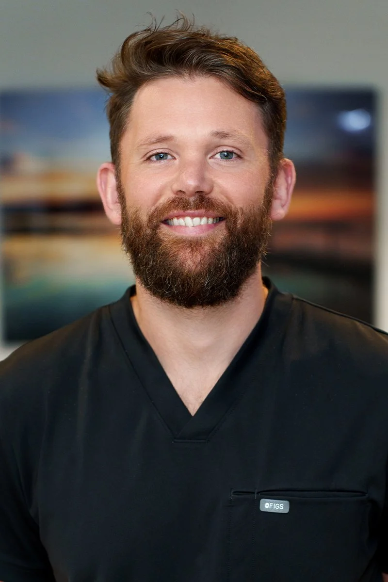 A man with a beard and short brown hair smiling, wearing a black medical scrub top, standing in front of a blurred background with a monitor and landscape image.